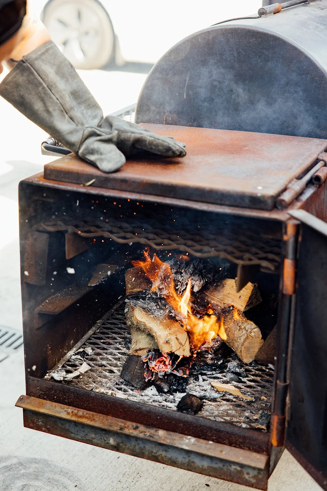 A person wearing a leather glove is adjusting a rusty outdoor smoker with burning wood inside.