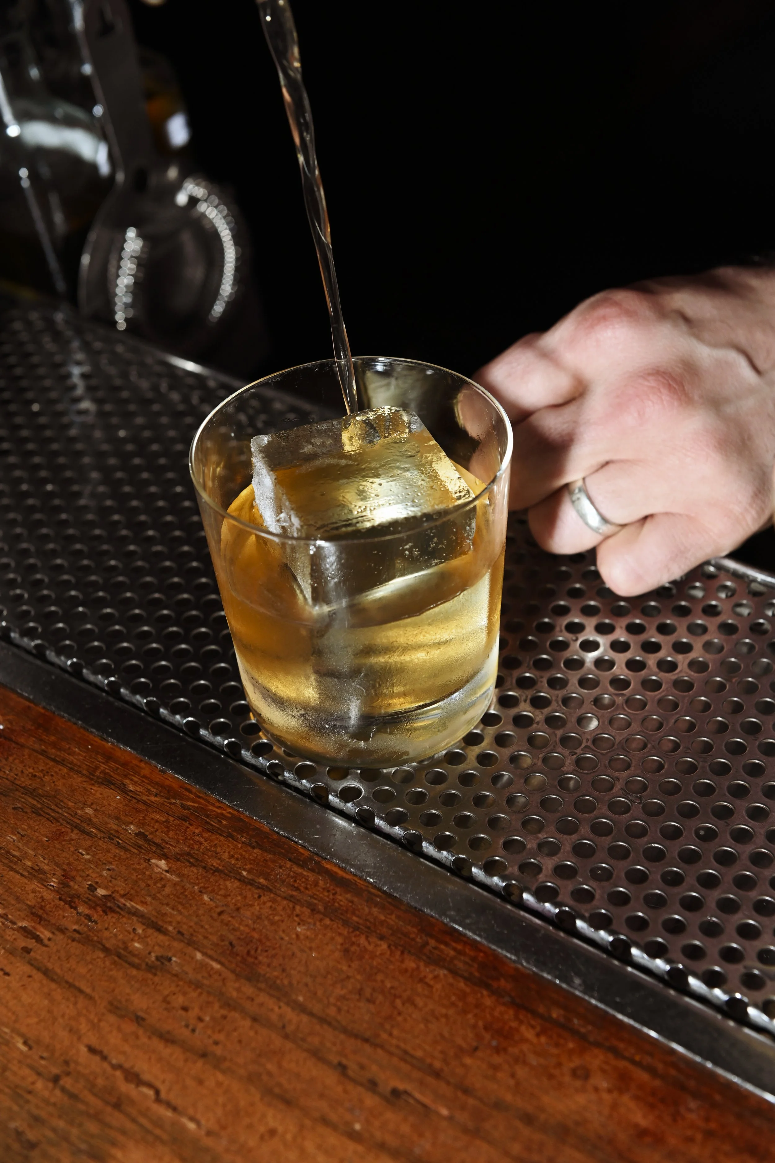A glass with a large ice cube, being filled with a golden-colored beverage, on a bar counter with a black rubber mat surface.