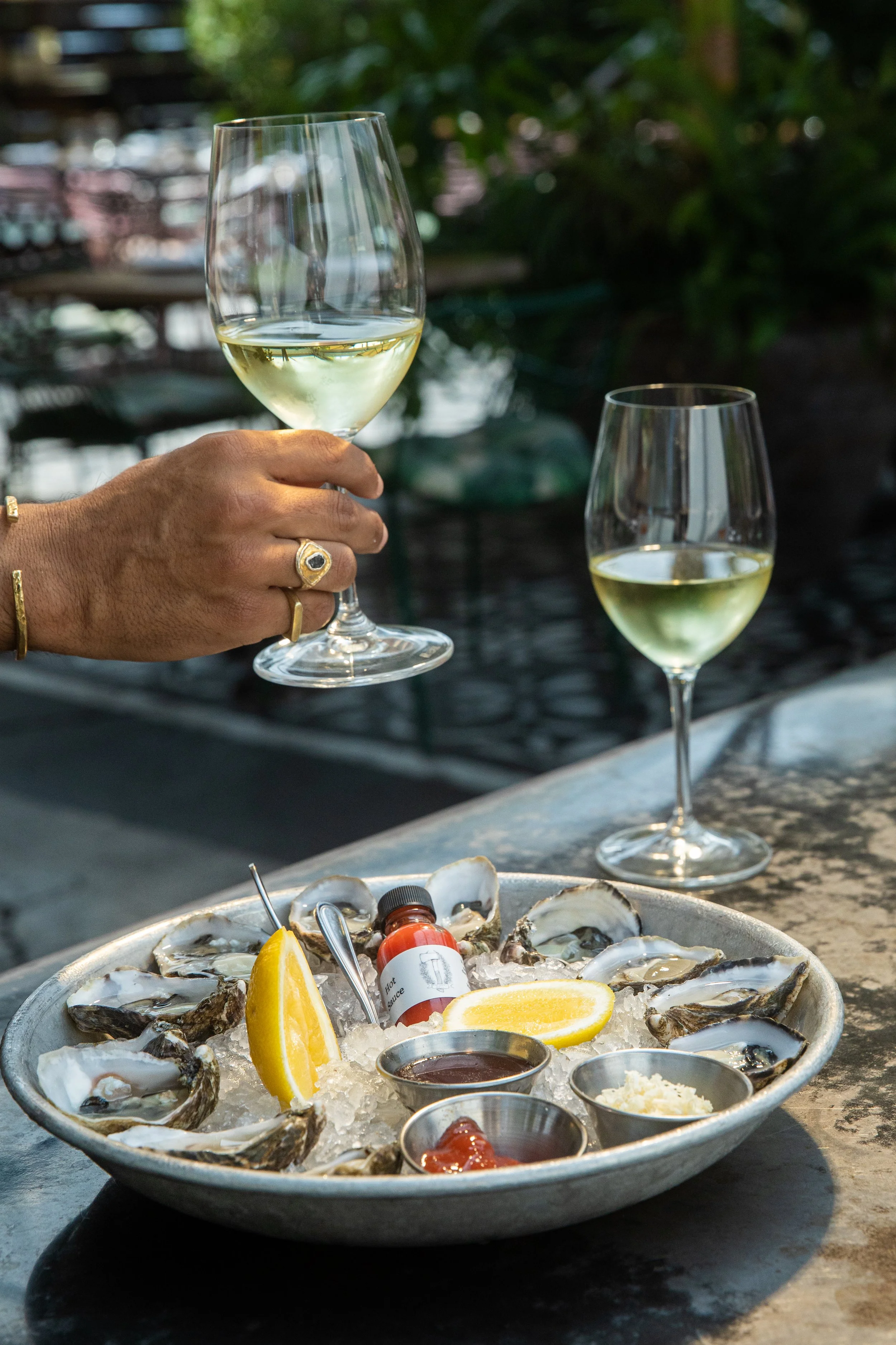 A person is holding a glass of white wine in front of a seafood platter on an outdoor table, with another glass of wine in the background, and oysters with lemon wedges on a bed of ice in the foreground.