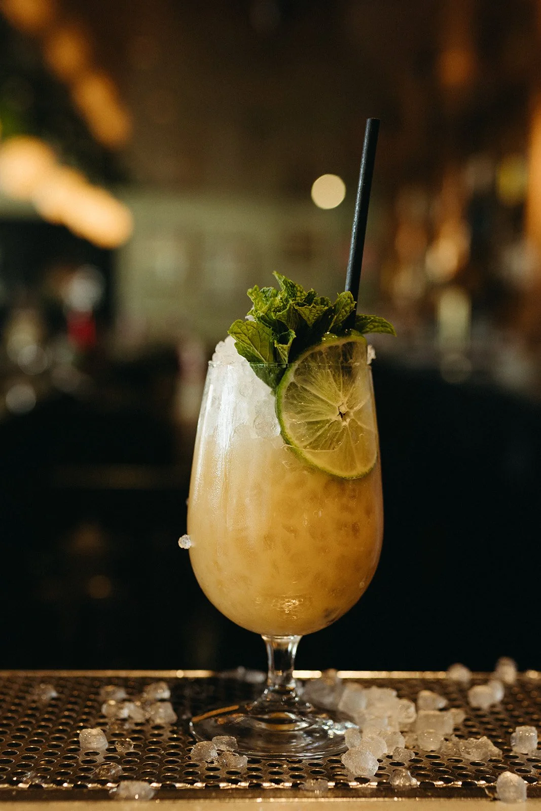 A tropical cocktail with a lime slice, mint leaves, ice, and a black straw in a tall glass, on a bar counter with ice scattered around.