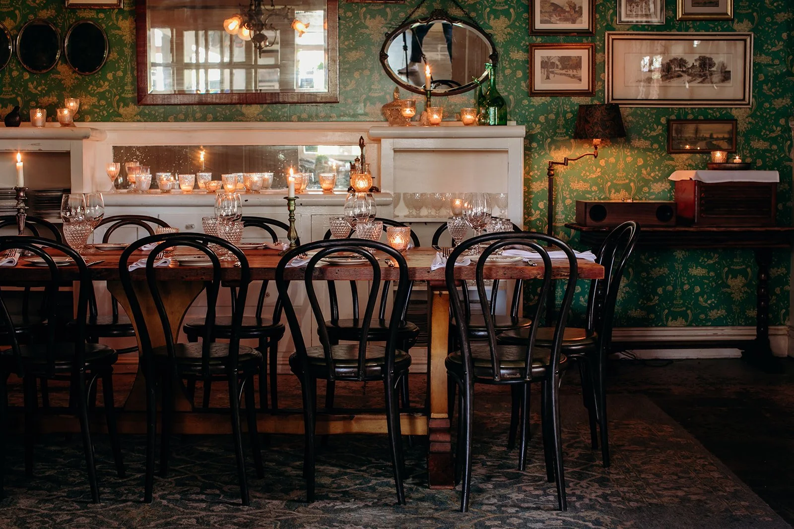 A dining room with a wooden table set with glasses and candles, surrounded by black chairs, and decorated with framed artwork on green wallpapered walls, featuring candles and a mirror.