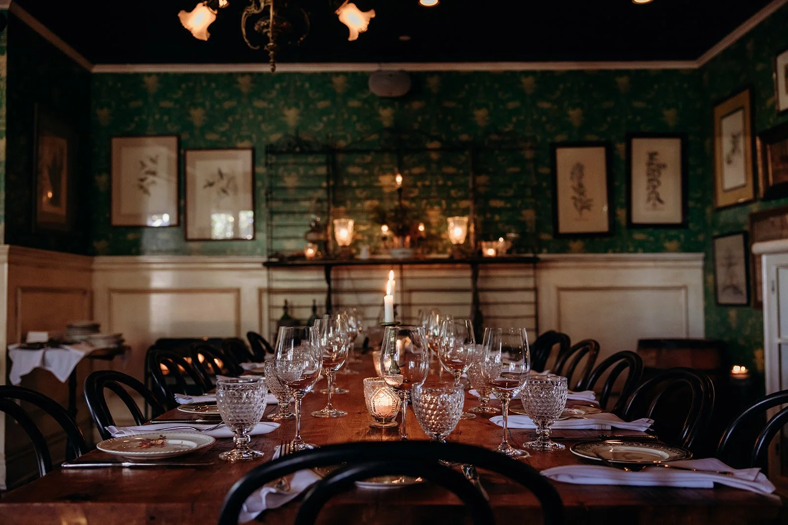 Dining table set with wine glasses, water glasses, plates, and napkins, decorated with candles in a dimly lit, vintage-style restaurant or dining room.