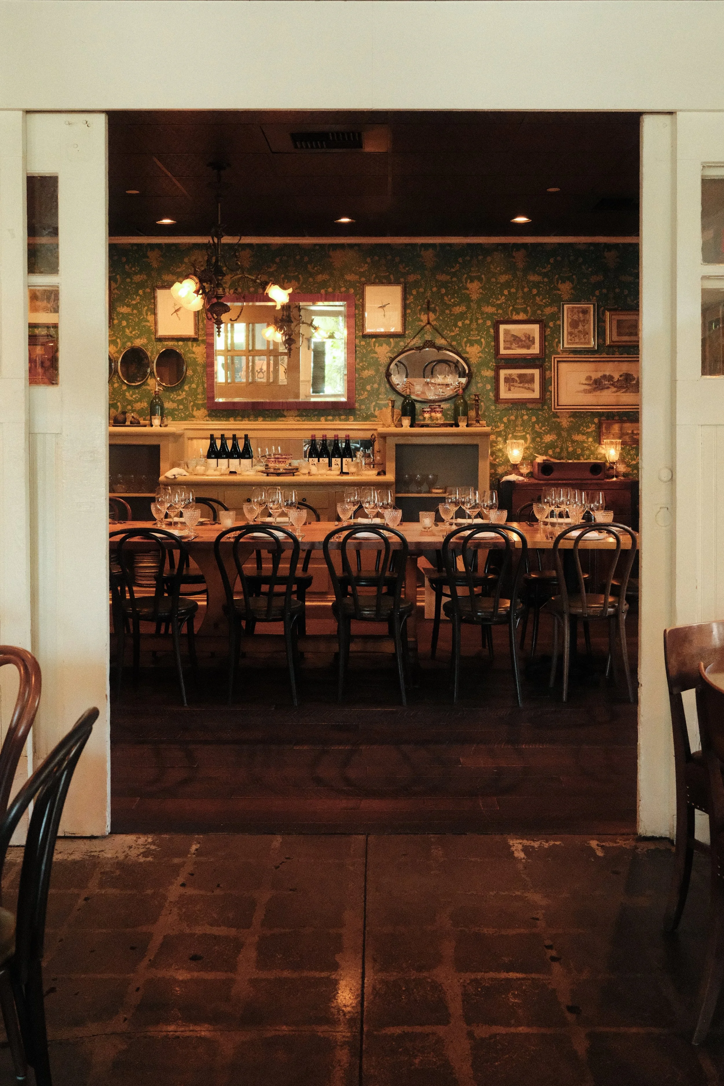 The photo shows a cozy dining room with a long table set with wine glasses and bottled water, surrounded by black wooden chairs. In the background, there is a decorative wall with framed art, a mirror, and bottles on a shelf. The room has warm lighting and a vintage, charming atmosphere.