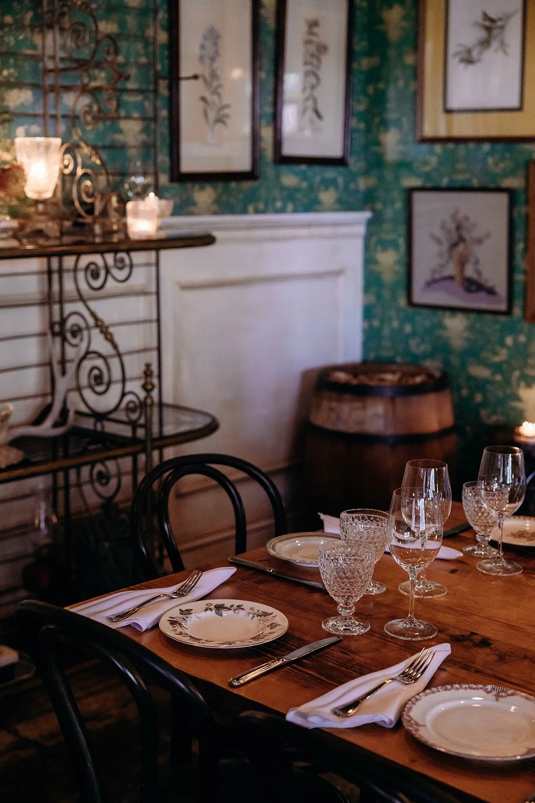 A cozy dining room with a wooden table set with plates, glasses, and silverware, decorated with white napkins. The background features framed artwork of trees on a green, textured wall, a barrel, and a metal shelf with candles and decorative items.
