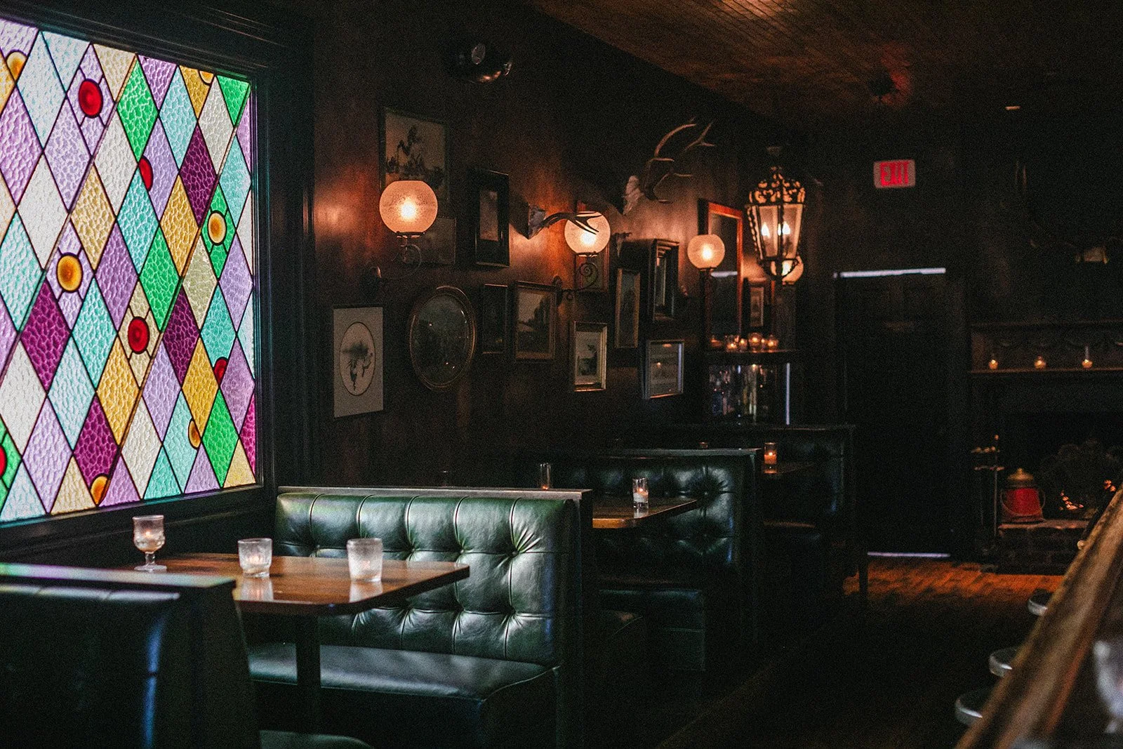 Dimly lit bar or restaurant interior with dark walls, velvet seating, framed pictures, and stained glass window with a colorful diamond pattern.