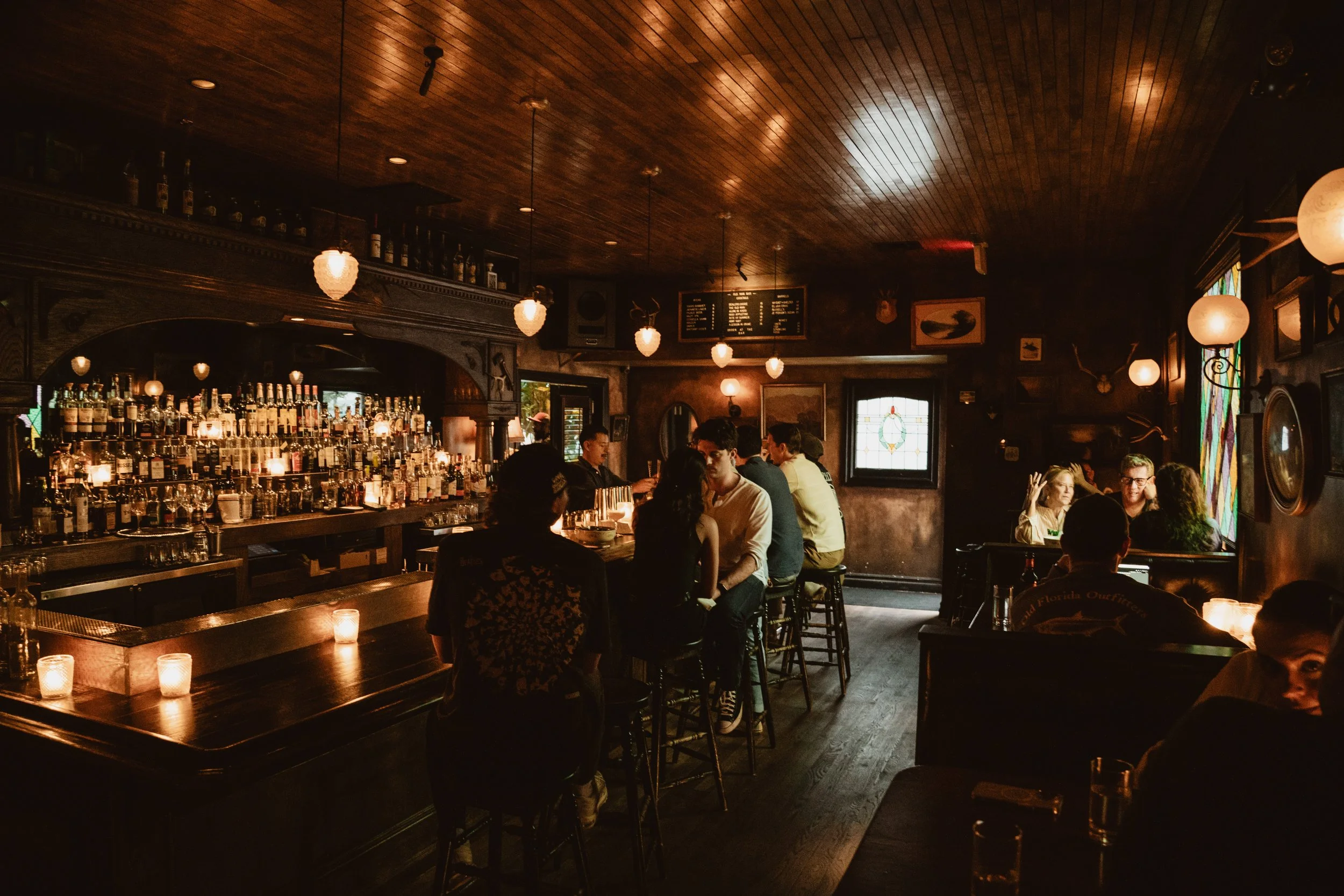 A cozy bar with a dark wooden ceiling and warm lighting. Several people are sitting and socializing at the bar and tables. The bar is stocked with bottles, and there are candles on the tables.