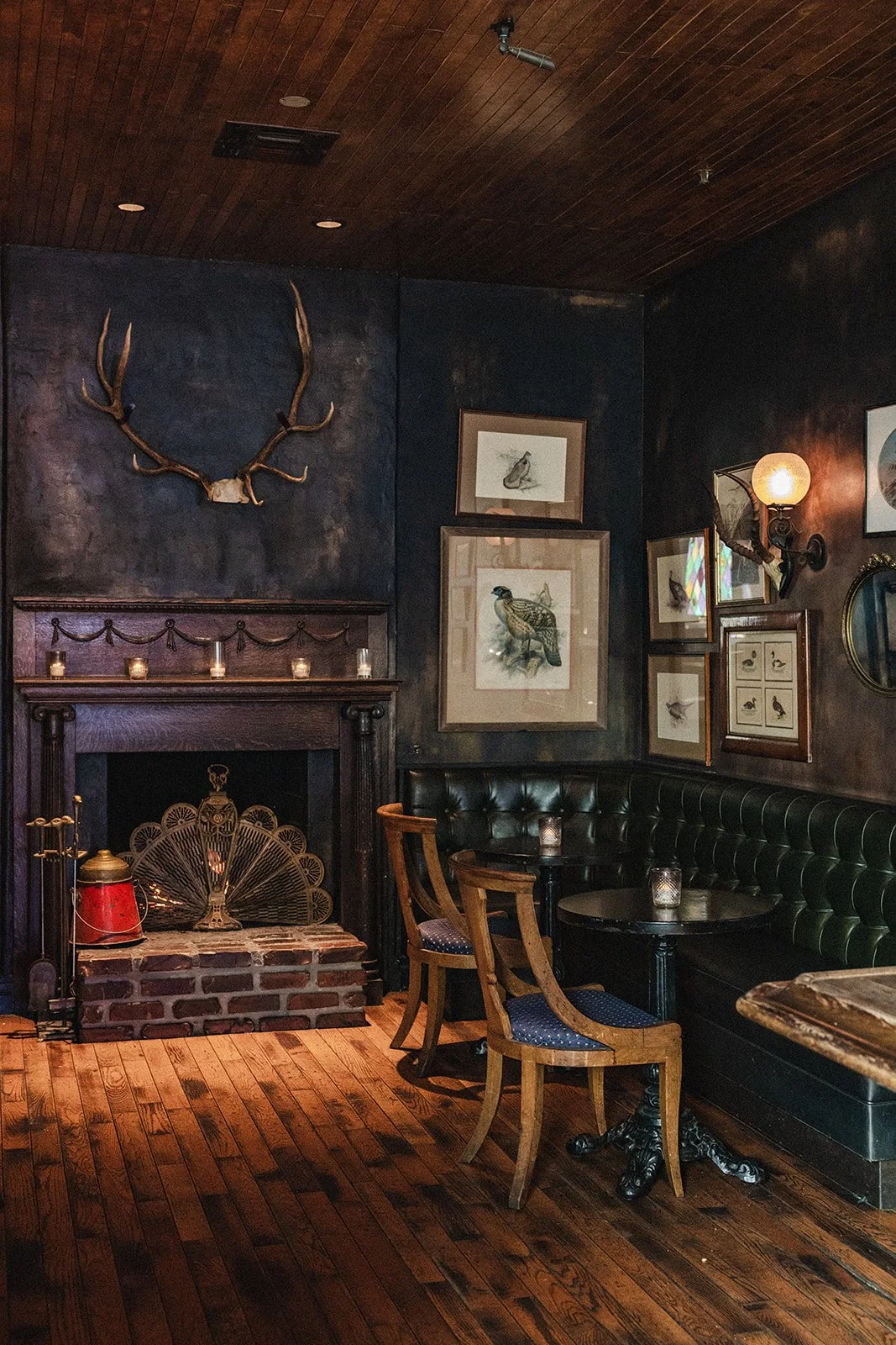 Interior of a cozy dimly lit room with dark walls, wooden ceiling, and floor. There is a brick fireplace with a decorative fan and candles on top. A mounted set of antlers is above the fireplace. Framed artwork and photographs of birds hang on the walls. A small round table with candles and two wooden chairs with blue cushioned seats are near a black leather banquette. An ornate wall sconce with a globe light is on the wall.