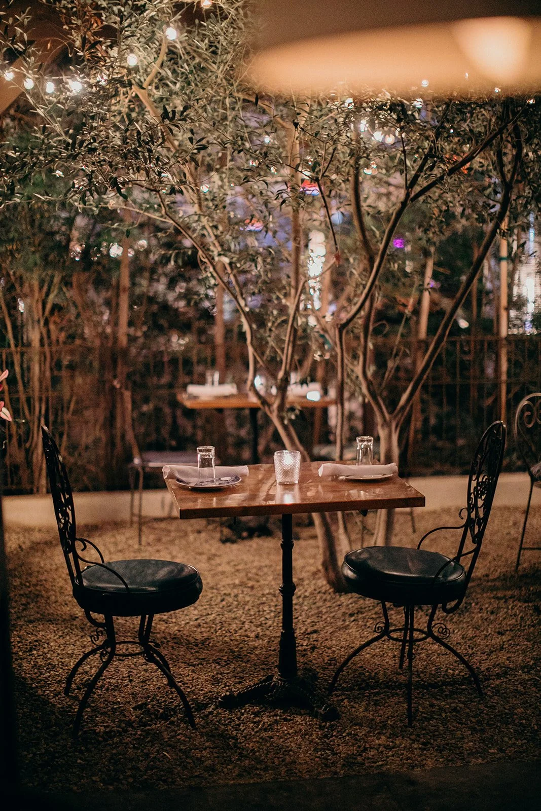A cozy outdoor dining setup with a wooden table set for two, surrounded by black wrought iron chairs, under a tree with string lights at night.