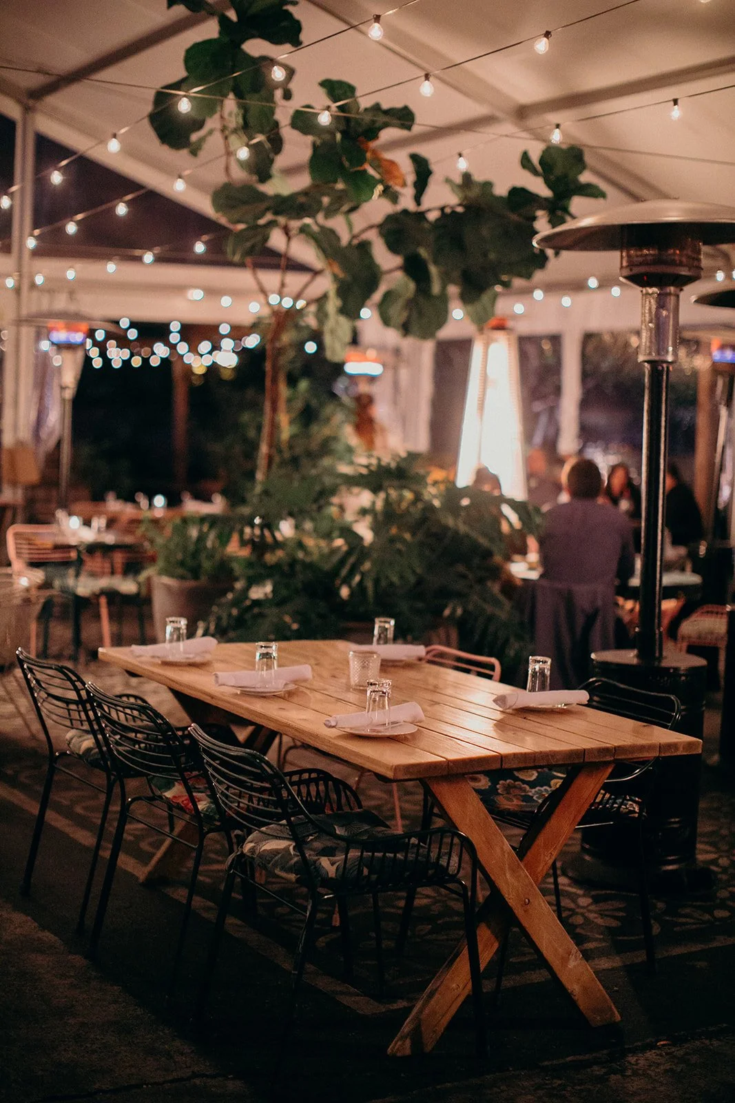 An outdoor dining area at night decorated with string lights, with a wooden table set with glasses and napkins, surrounded by black chairs, and a heater standing nearby.