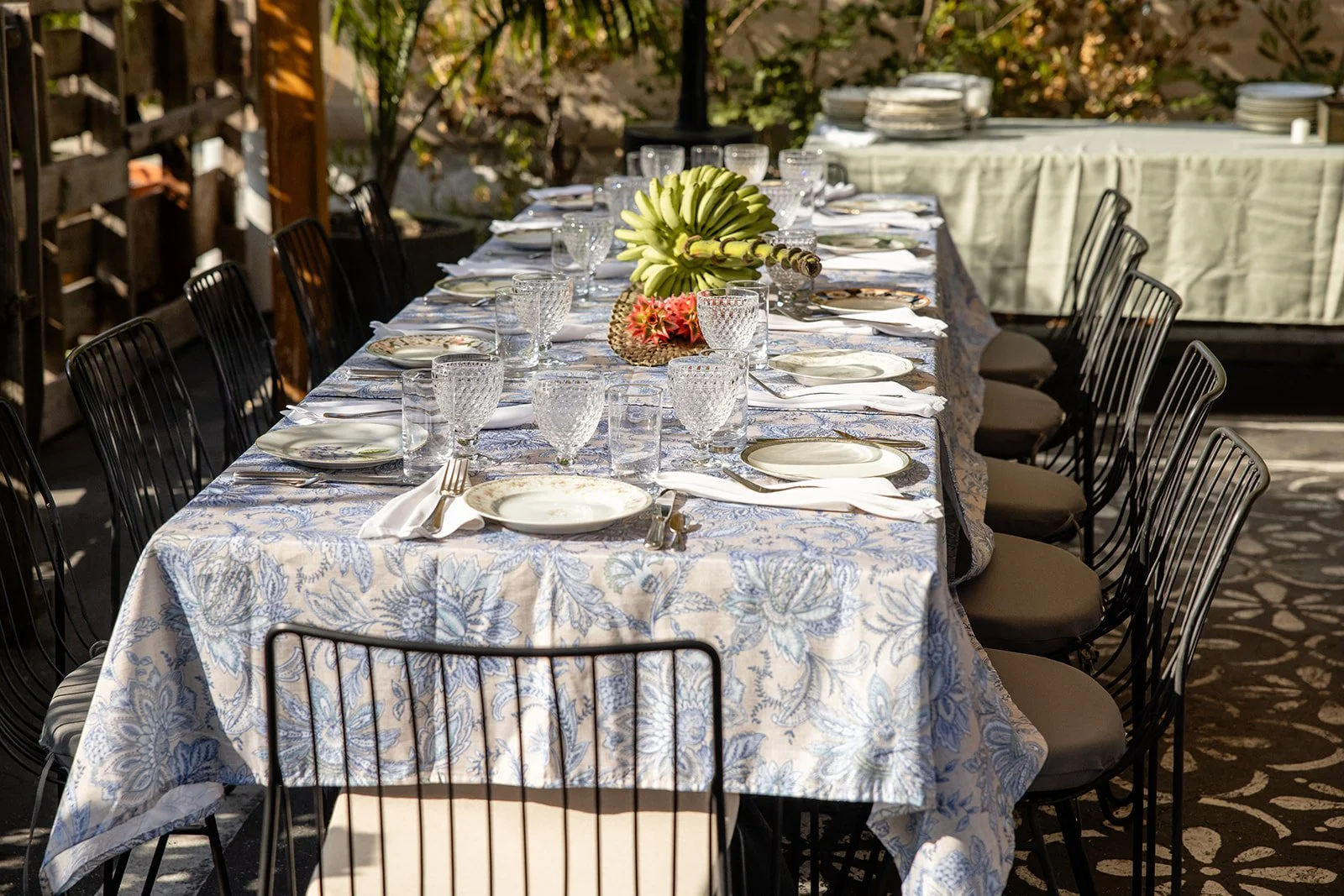 A long outdoor dining table set for a meal, with plates, glassware, and silverware, covered with a floral tablecloth and decorated with a banana and pineapple centerpiece. Chairs are arranged around the table, and stacks of plates are visible in the background.