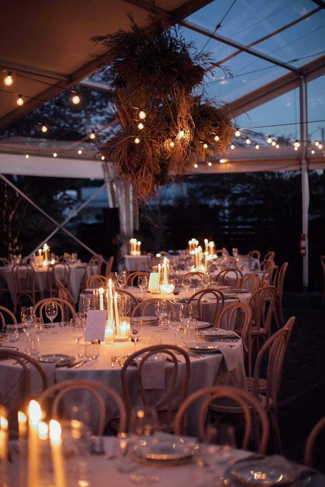 Elegant outdoor dining setup at dusk with round tables covered in white tablecloths, decorated with candles and glassware, under a tent with hanging string lights and a large floral and greenery arrangement overhead.