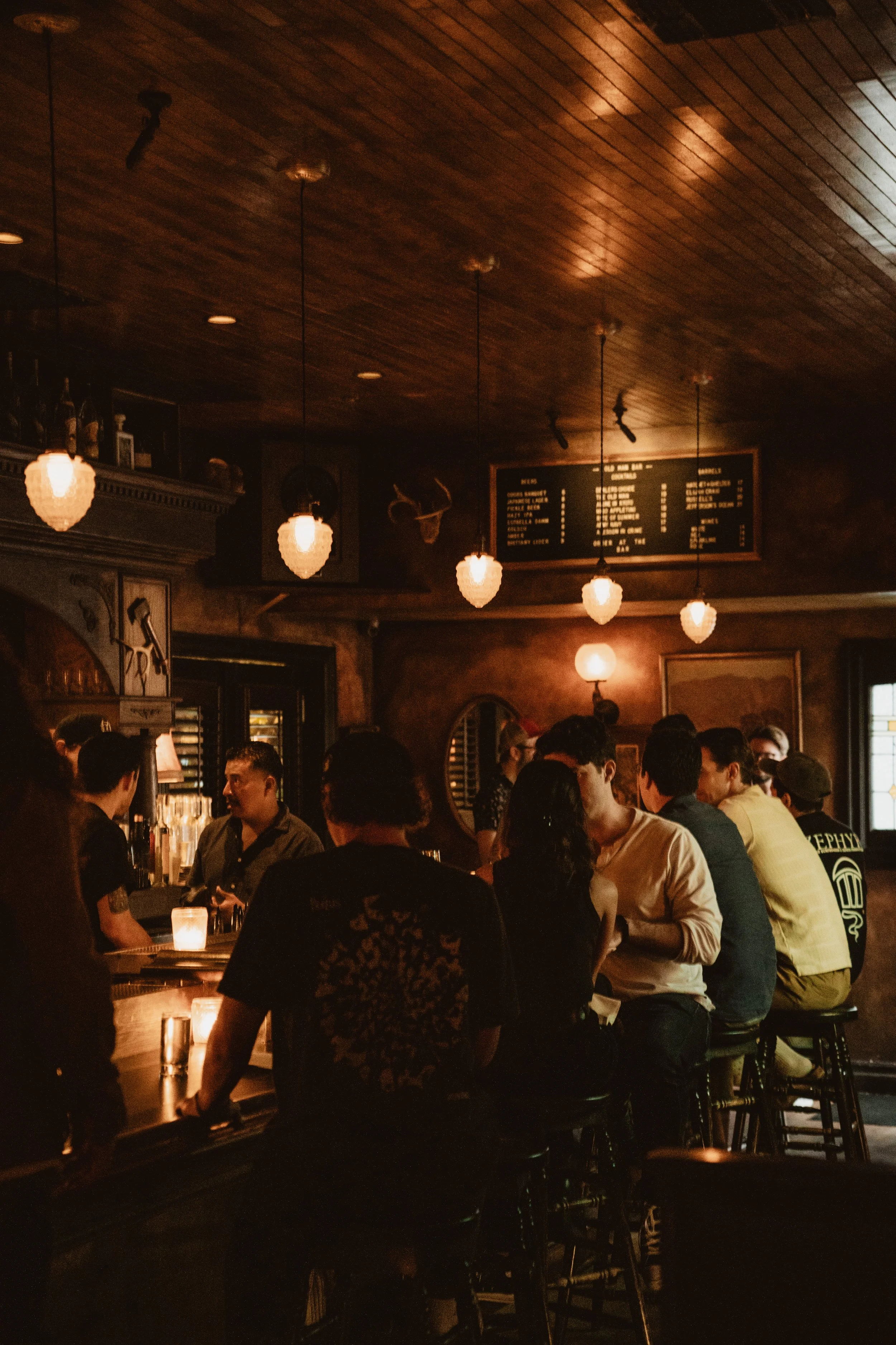 A dimly lit bar or pub with a wooden ceiling and hanging pendant lights, crowded with people socializing around the bar counter.