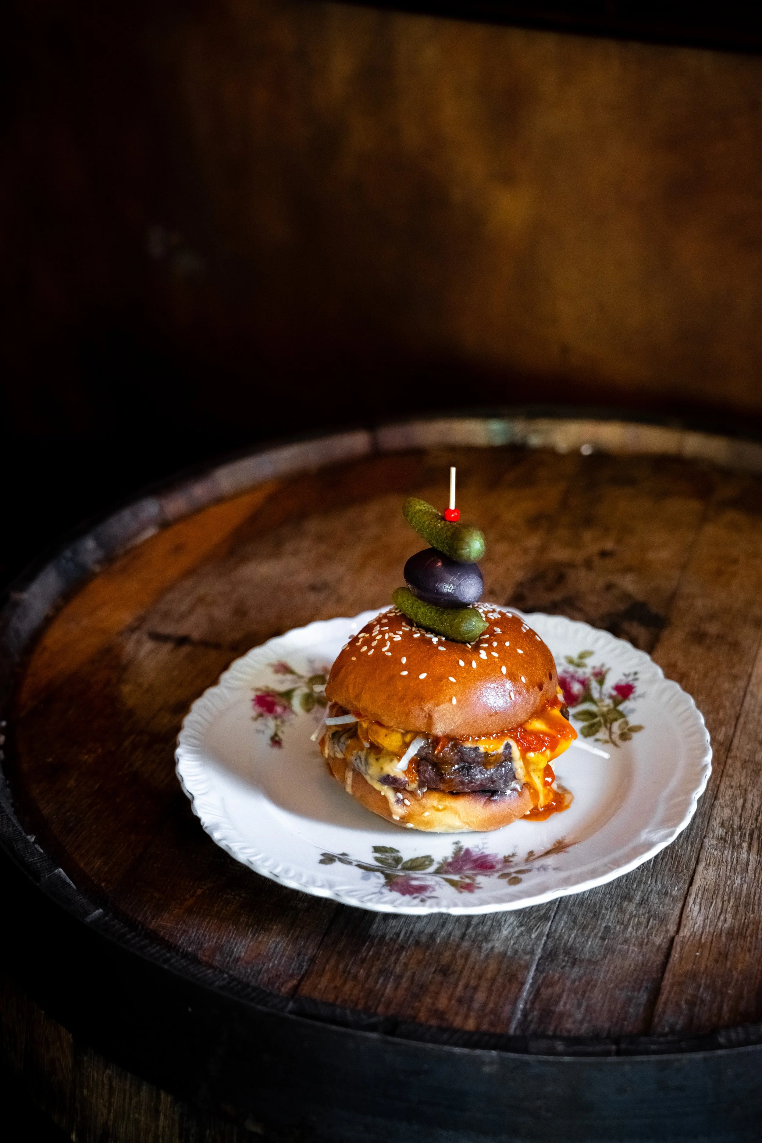 A hamburger with a sesame seed bun, cheese, and toppings, topped with an olive, a small pickle, and a toothpick with a red bead, served on a decorative plate with floral patterns.