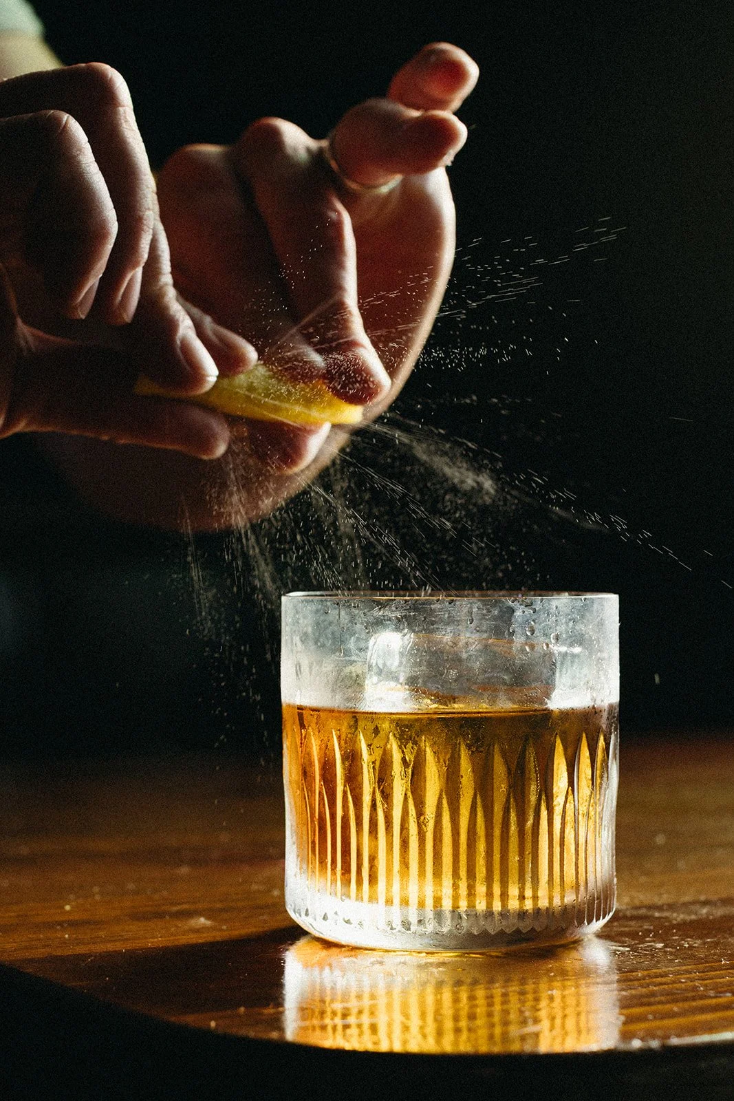 Hand squeezing a lemon slice into a glass of whiskey or similar amber-colored liquor on a dark background.
