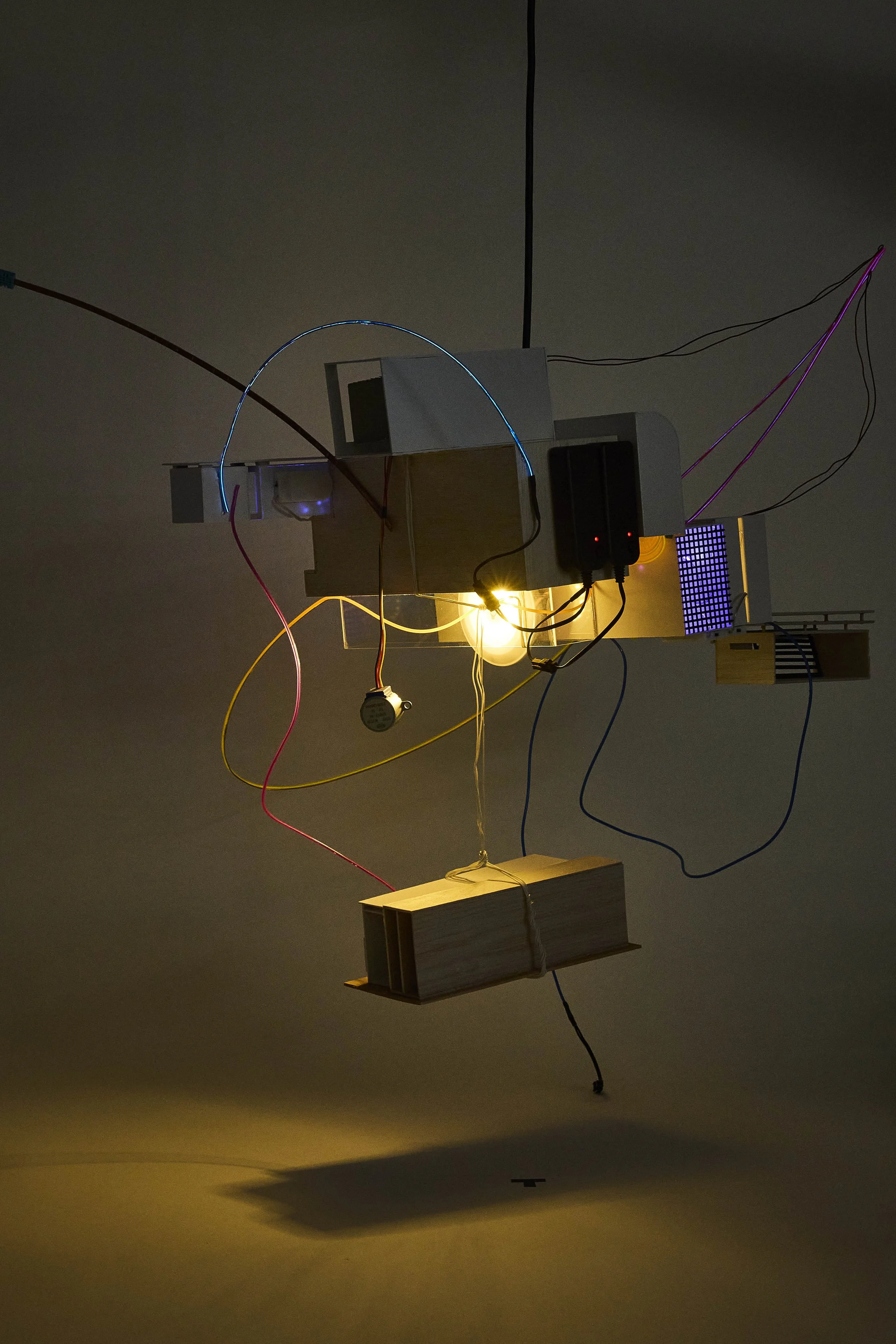 An abstract artistic light installation hanging from the ceiling, consisting of various rectangular boxes, wires, and illuminated elements, casting shadows on the wall below.
