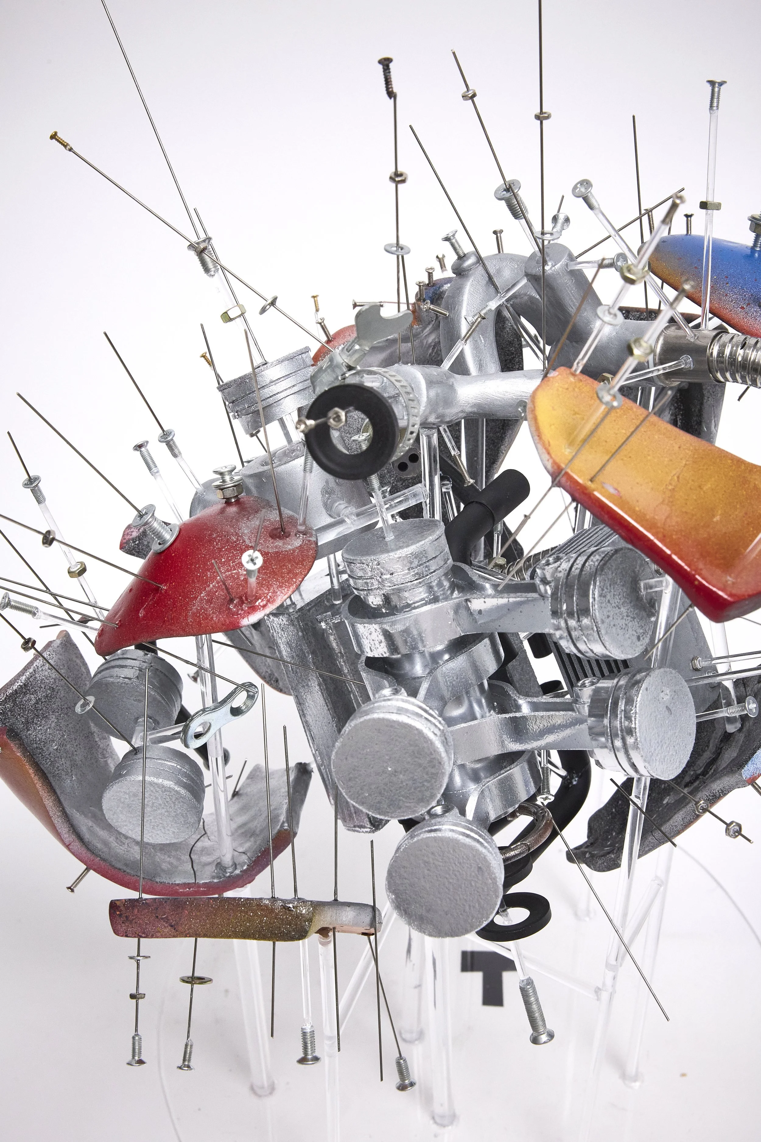 An abstract sculpture made of various metallic and painted parts, with numerous nails and screws protruding in different directions, creating a chaotic, three-dimensional composition.