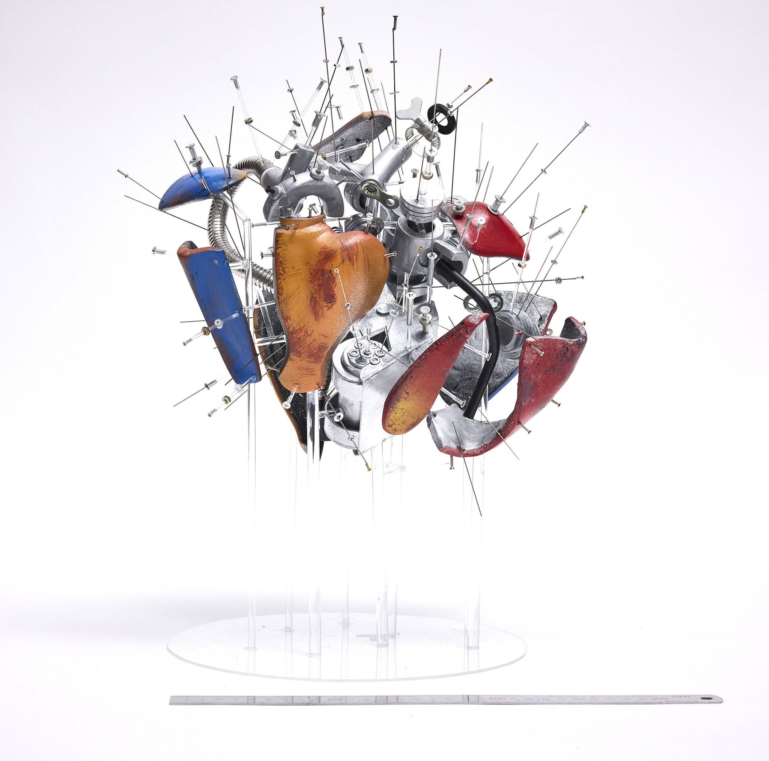 An artistic sculpture resembling a human heart made from various metal parts, with colorful painted sections and numerous nails protruding from it.
