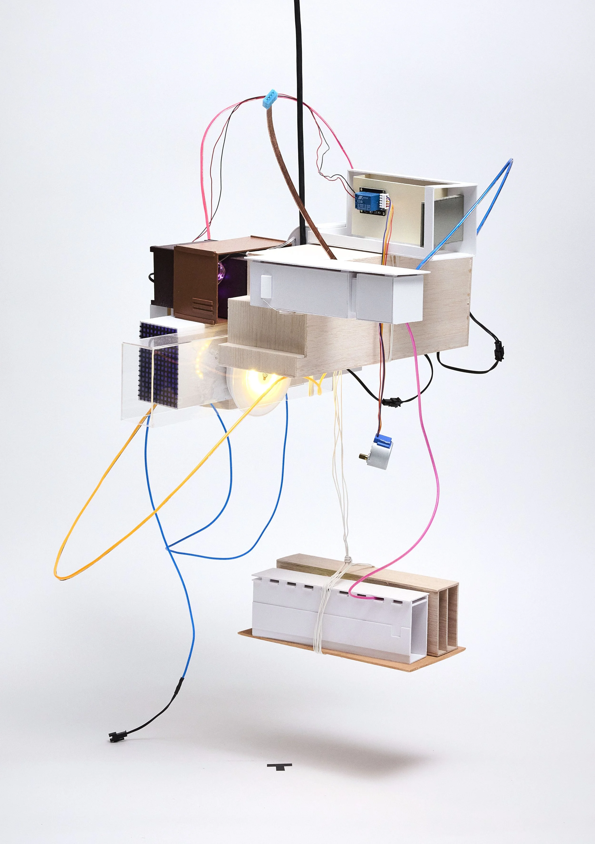 A contemporary art installation composed of various wooden and plastic geometric shapes, wires, and electronic components, suspended in midair against a plain white background.