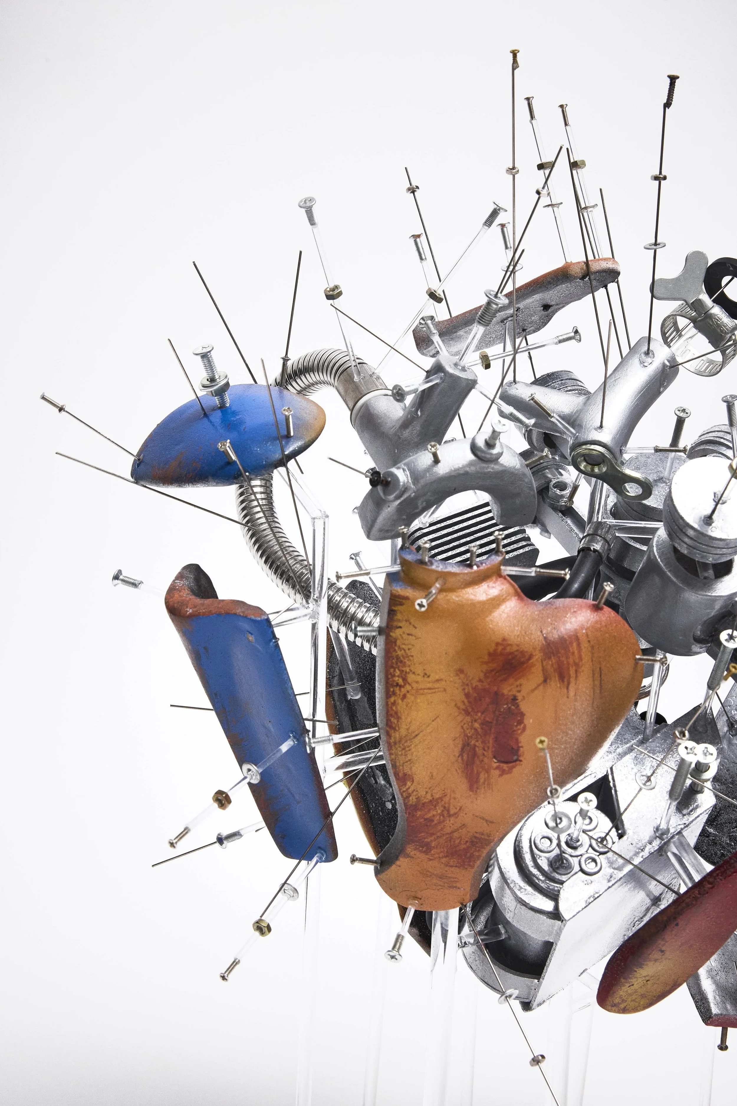 Abstract sculpture made of metal parts, bolts, and screws, with various colors and textured surfaces, mounted on a white background.