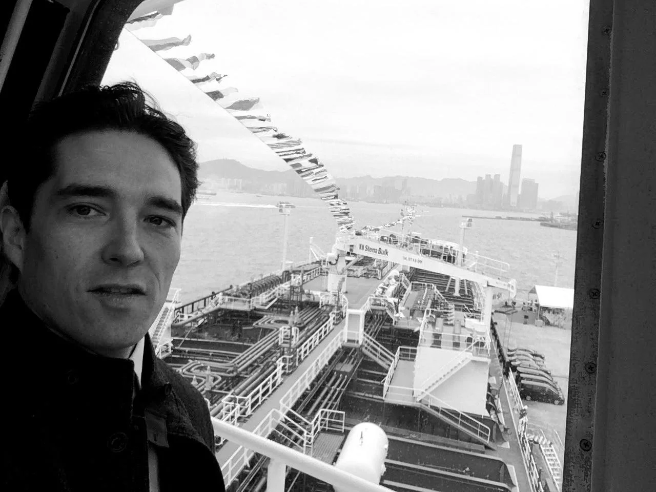 A young man taking a selfie inside a ferry with a view of the water and city skyline in the background.