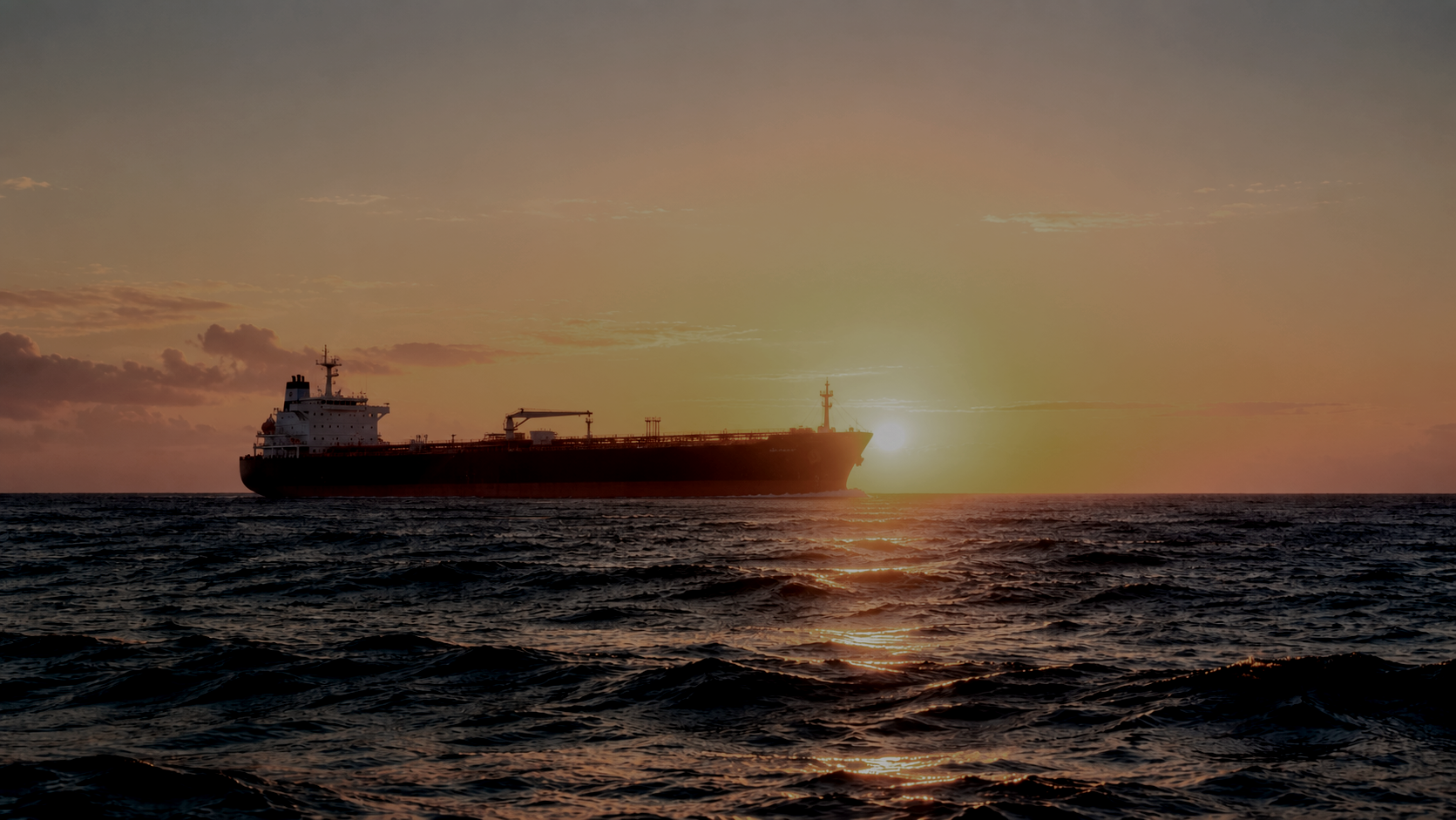 A large cargo ship sailing on the ocean during sunset with the sun low on the horizon, casting a glow and creating a silhouette of the ship against the sky.