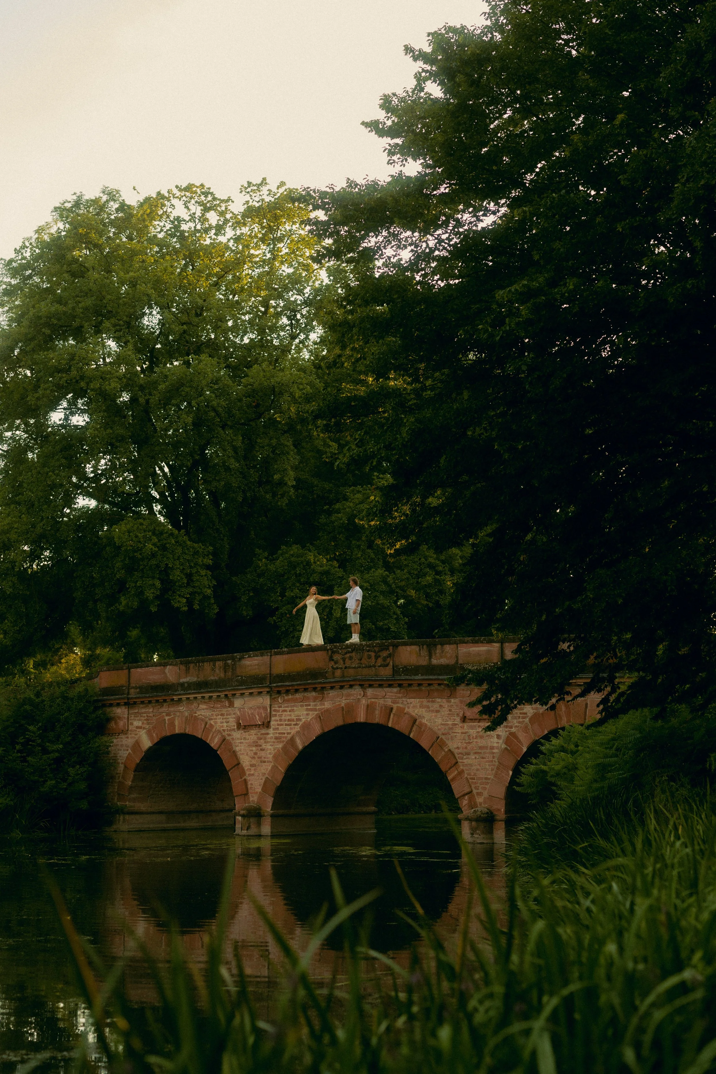 Ein Paar tanzt auf einer Brücke über einem Fluss, umgeben von großen Bäumen, bei Sonnenuntergang.
