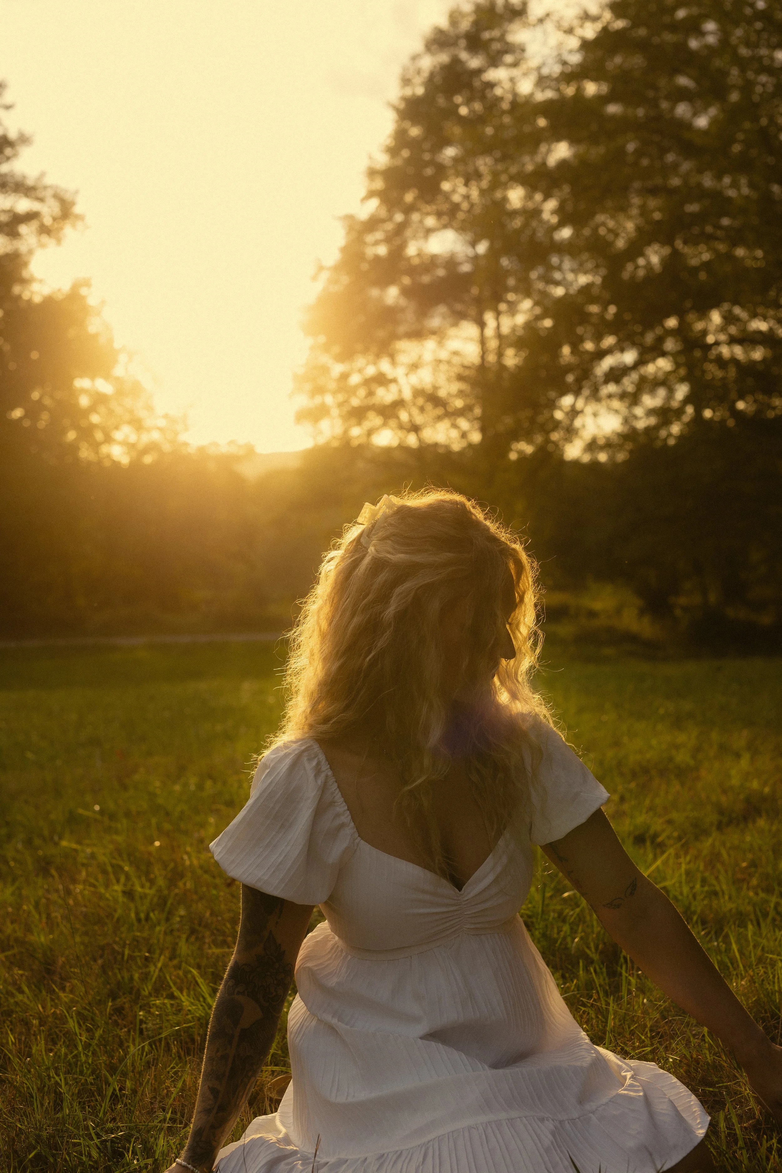 Eine Frau mit langen, blonden Haaren sitzt im Gras bei Sonnenuntergang in einem Park oder Wiese. Sie trägt ein weißes, lockeres Kleid und hat Tattoos auf den Armen. Im Hintergrund sind Bäume und die Sonne, die den Himmel orange färbt, sichtbar.