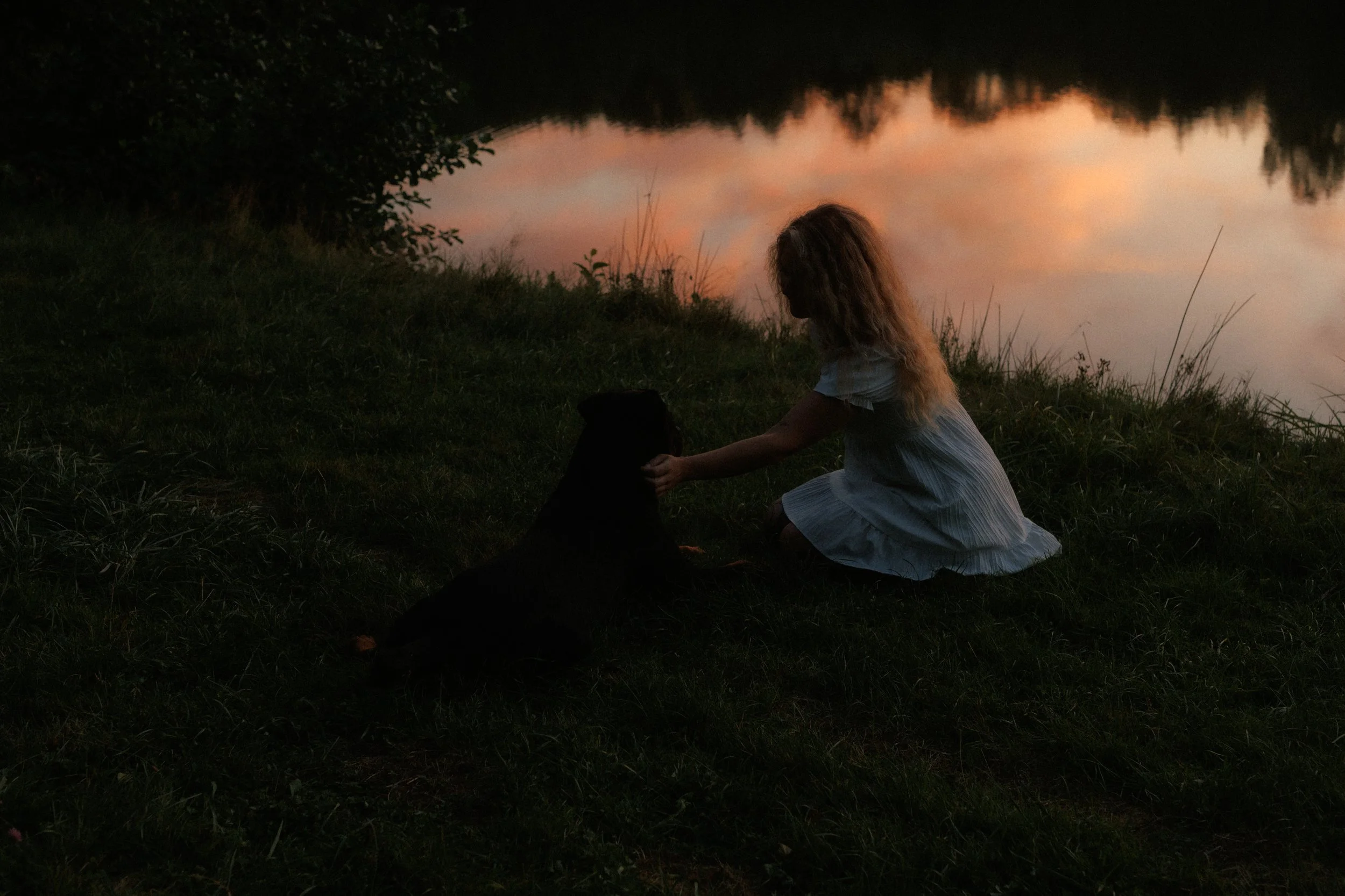 Eine junge Frau mit langen, lockigen Haaren, die in einem weißen Kleid auf einer Wiese neben einem See sitzt. Es ist Abend oder Nacht, mit einem orangefarbenen Himmel und reflektiertem Wasser im Hintergrund.