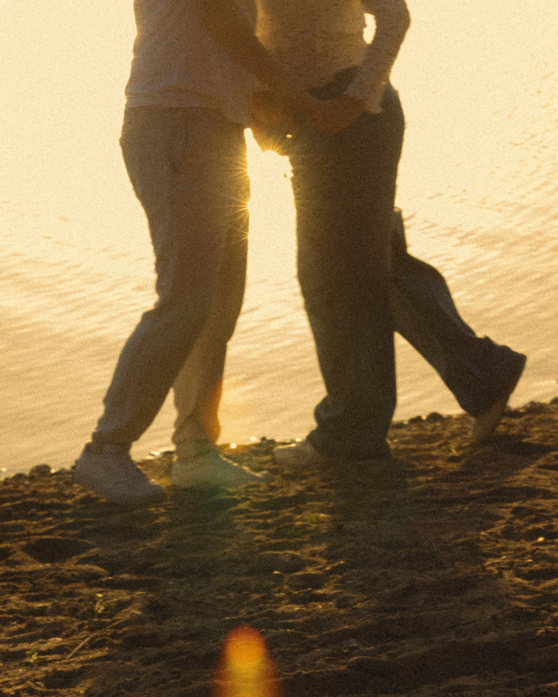 Schatten zweier Menschen, die sich an einem sonnigen Strand bei Sonnenuntergang oder Sonnenaufgang umarmen, mit sandigem Boden und einem kleinen Stein im Vordergrund.