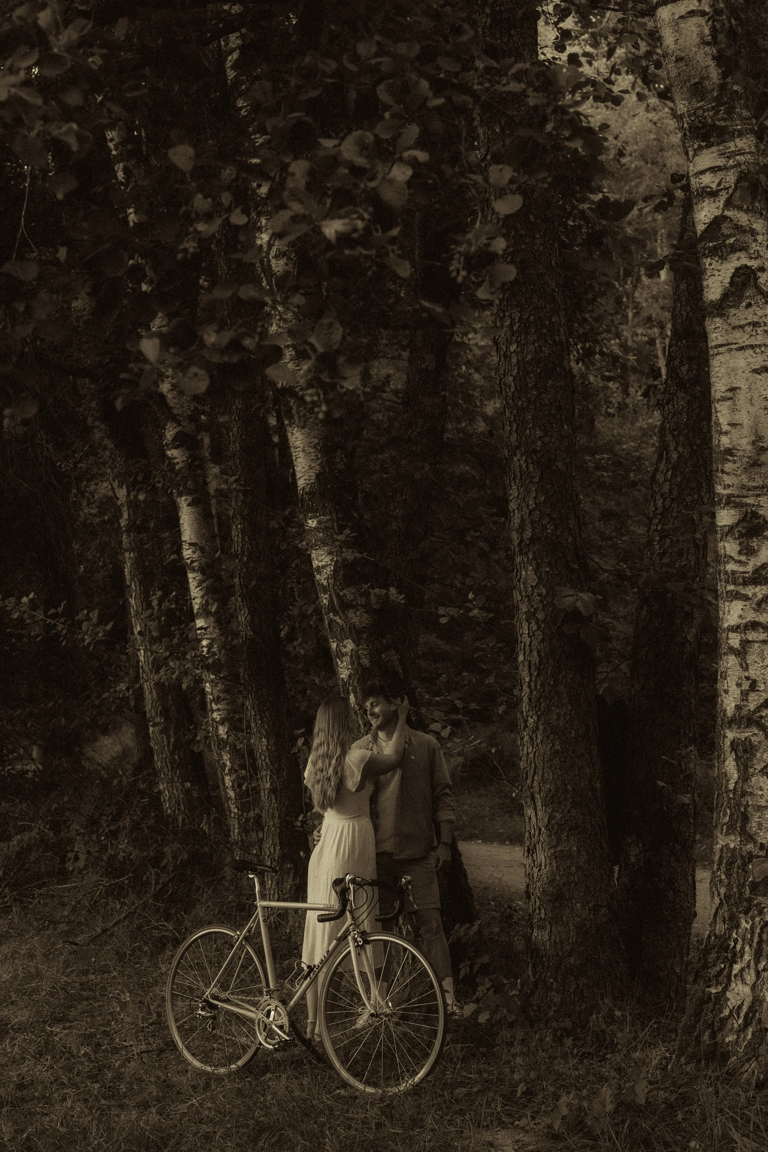 Zwei Menschen stehen sich im Wald gegenüber, eine Frau mit langem, gewelltem Haar in einem weißen Kleid, ein Mann in einem dunklen Anzug. Es befindet sich ein Fahrrad in der Nähe im Gras.