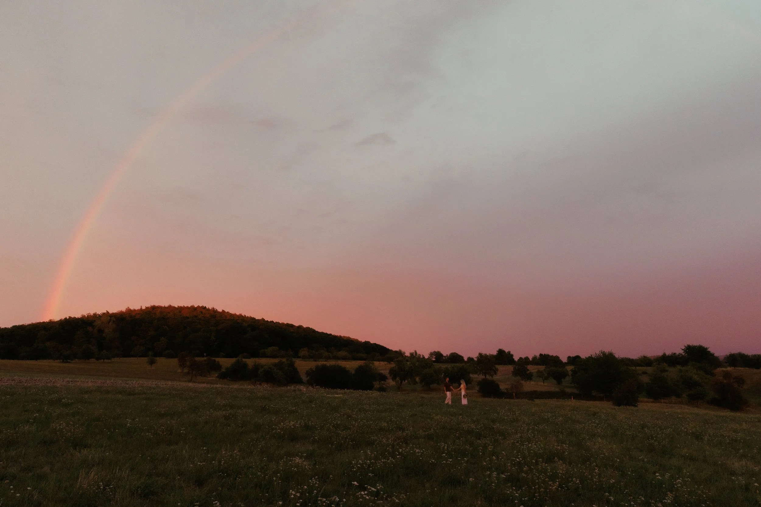 Ein Regenbogen erstreckt sich über einen Hügel mit Bäumen bei Sonnenuntergang, im Vordergrund sind zwei Menschen, die Hand in Hand im Feld stehen.