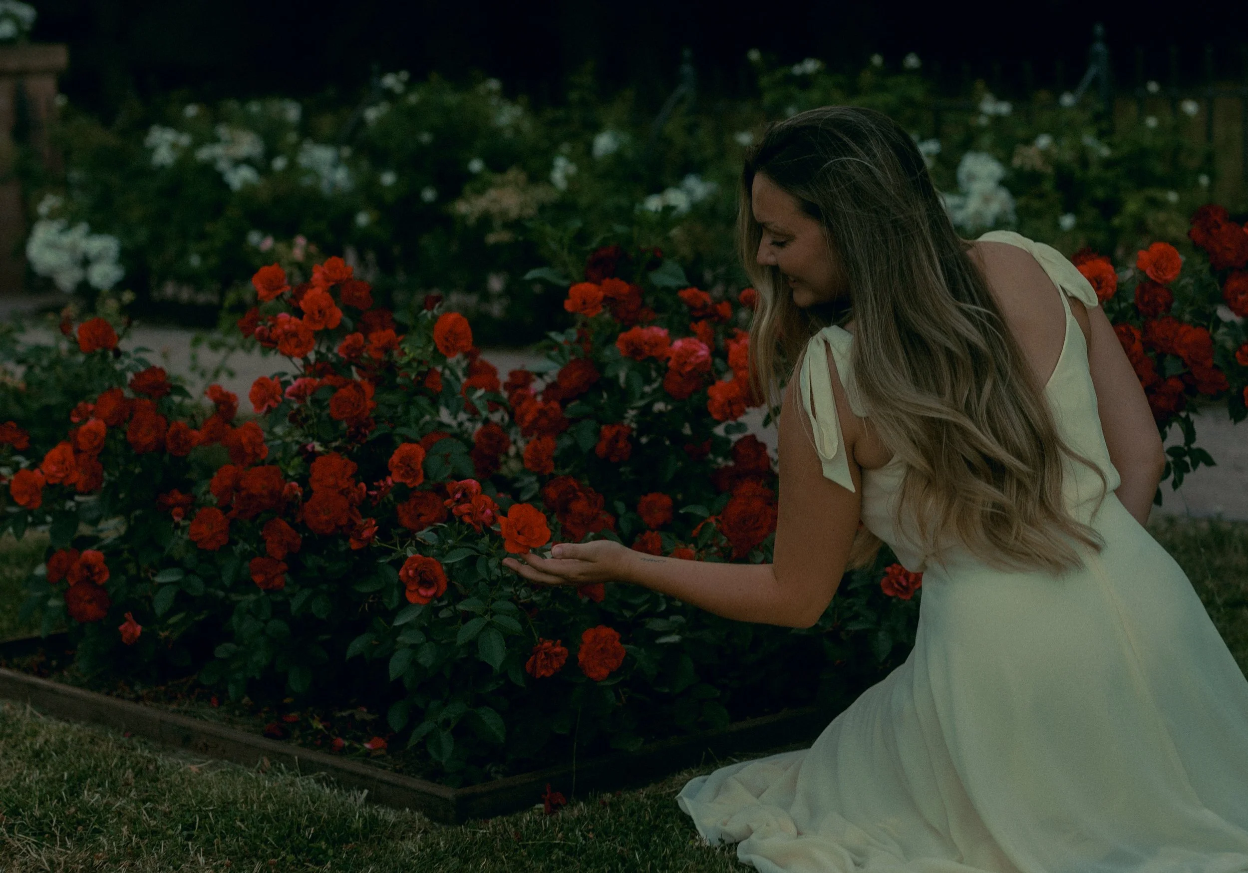 Eine Frau in einem weißen Kleid sitzt im Garten vor einem Blumenbeet mit roten und weißen Blumen, bei Nacht.