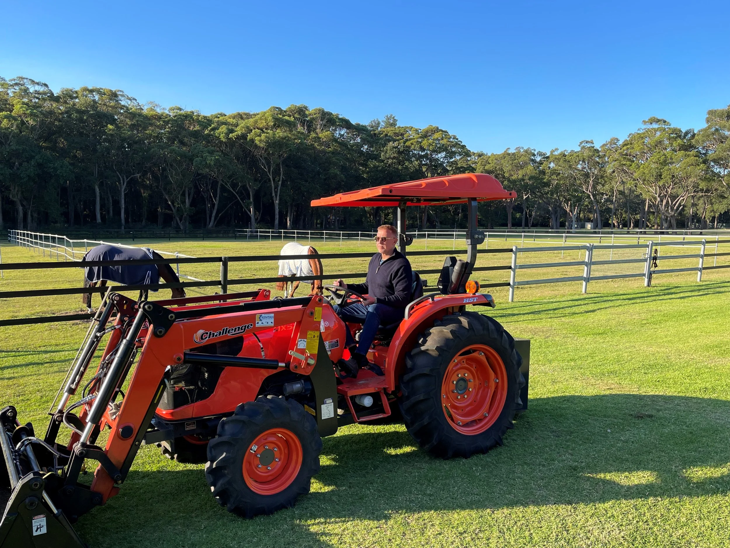 A man driving an orange compact tractor with a black and red attachment in a grassy area with a white fence and trees in the background.
