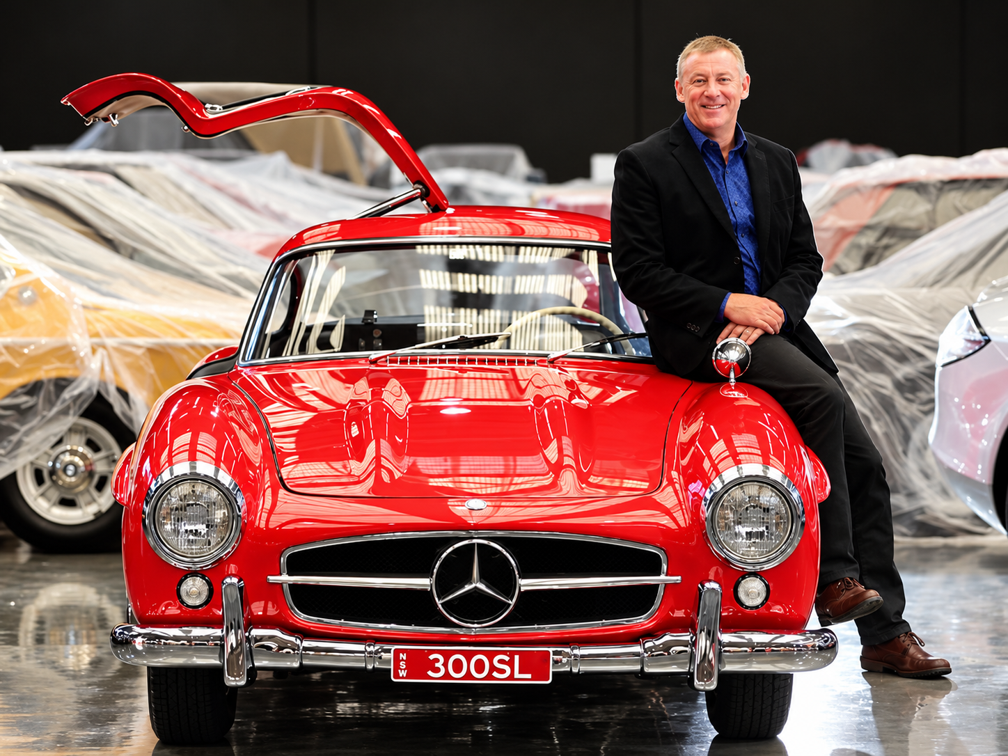 A man in a black blazer and blue shirt sitting on the hood of a red vintage Mercedes-Benz 300SL sports car in a showroom with covered cars in the background.