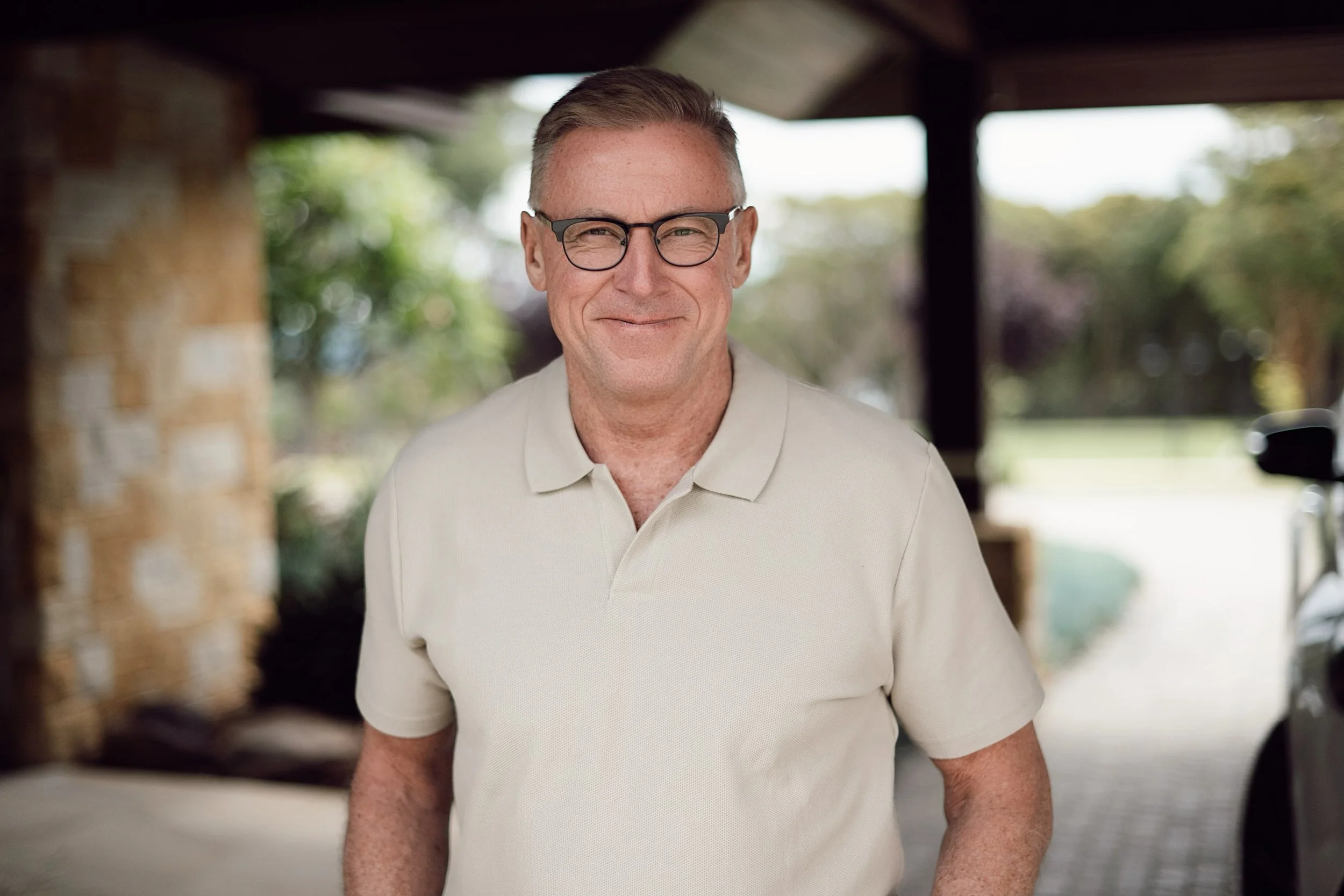 A middle-aged man with short gray hair, wearing glasses and a light-colored polo shirt, smiling in an outdoor setting with blurred greenery in the background.