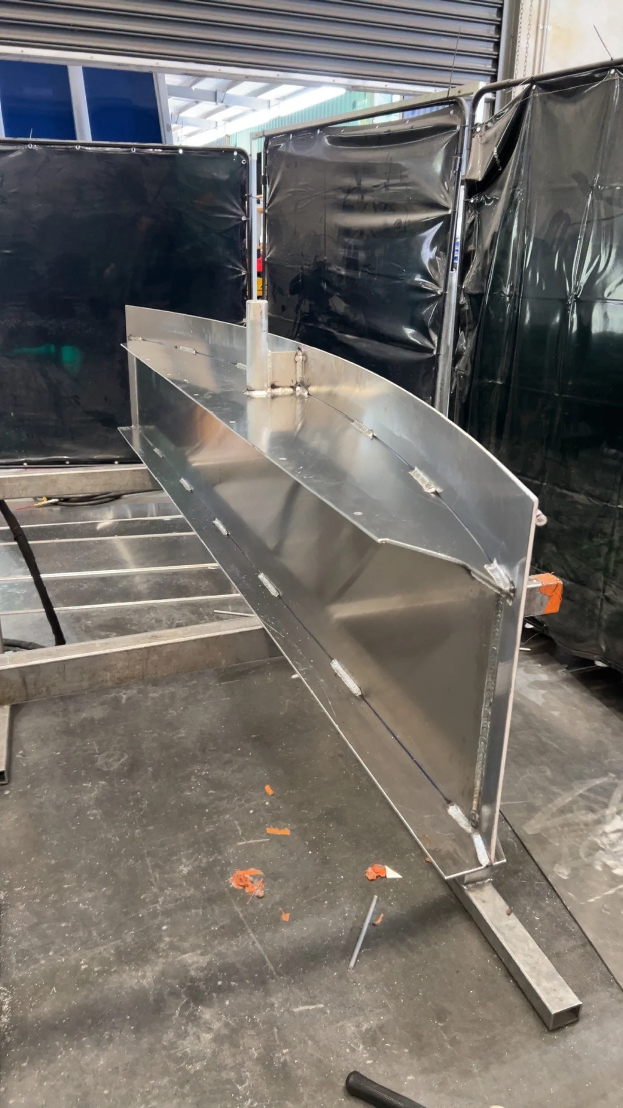 Metal boat hull frame under construction in a workshop, with tools and debris on a concrete floor.