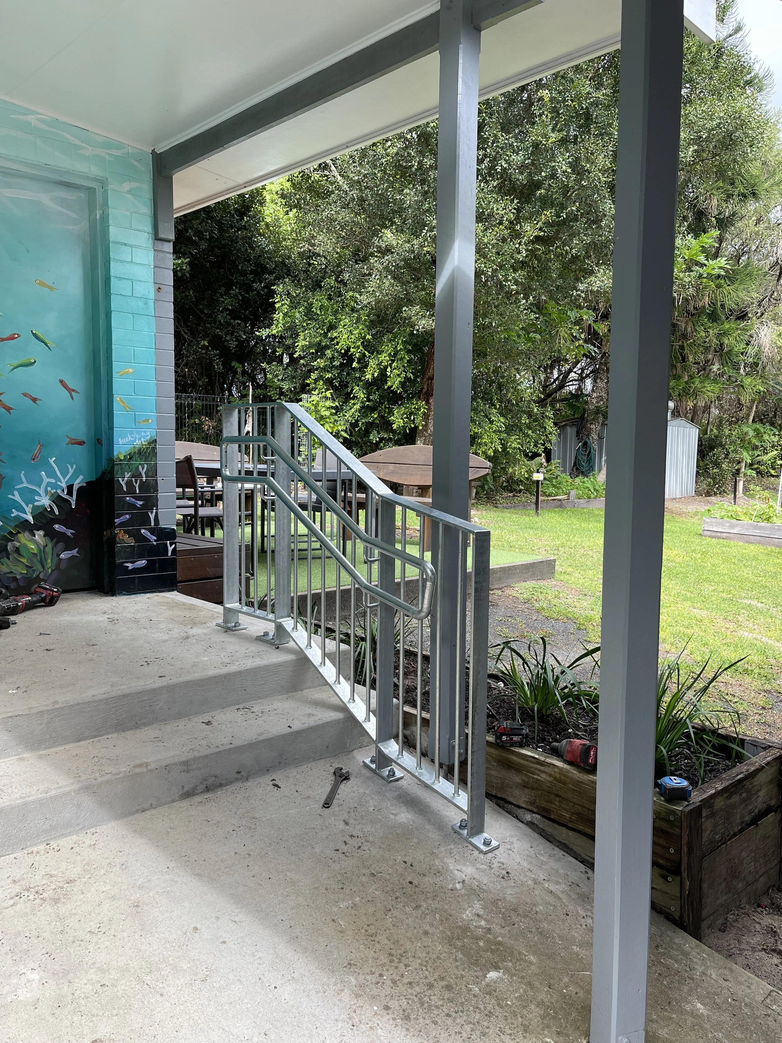 Outdoor patio area under construction with metal railings, concrete steps, a table and chairs, garden, trees, and a small shed in the background.