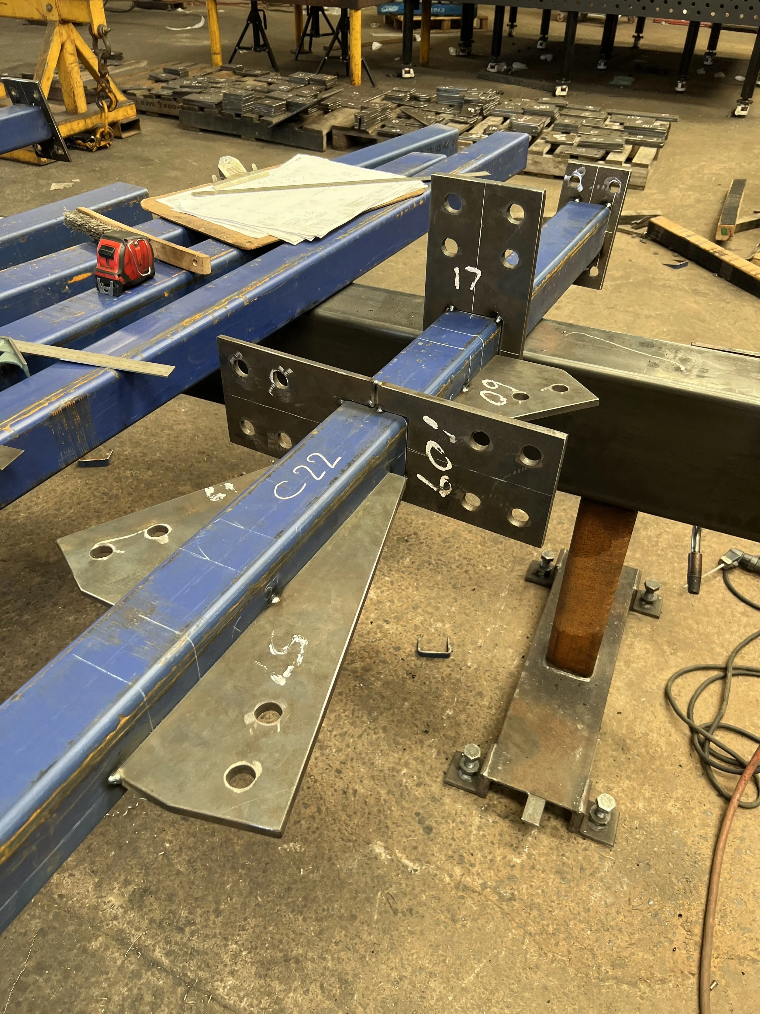 A construction or workshop area with metal beams and metal plates, some with drilled holes, being assembled or welded. Tools and papers are scattered on top of the beams, including a measuring tape and a carpenter's square. There are pallets and stacks of metal pieces in the background.
