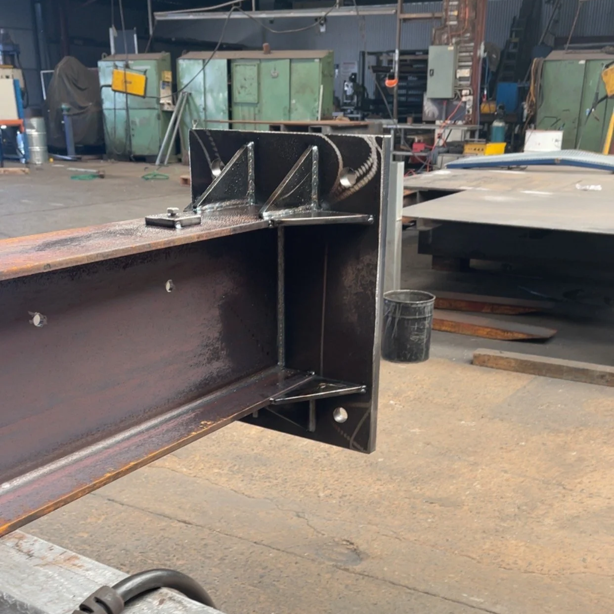 A steel I-beam with welded brackets and chain attachments inside a workshop or factory setting.