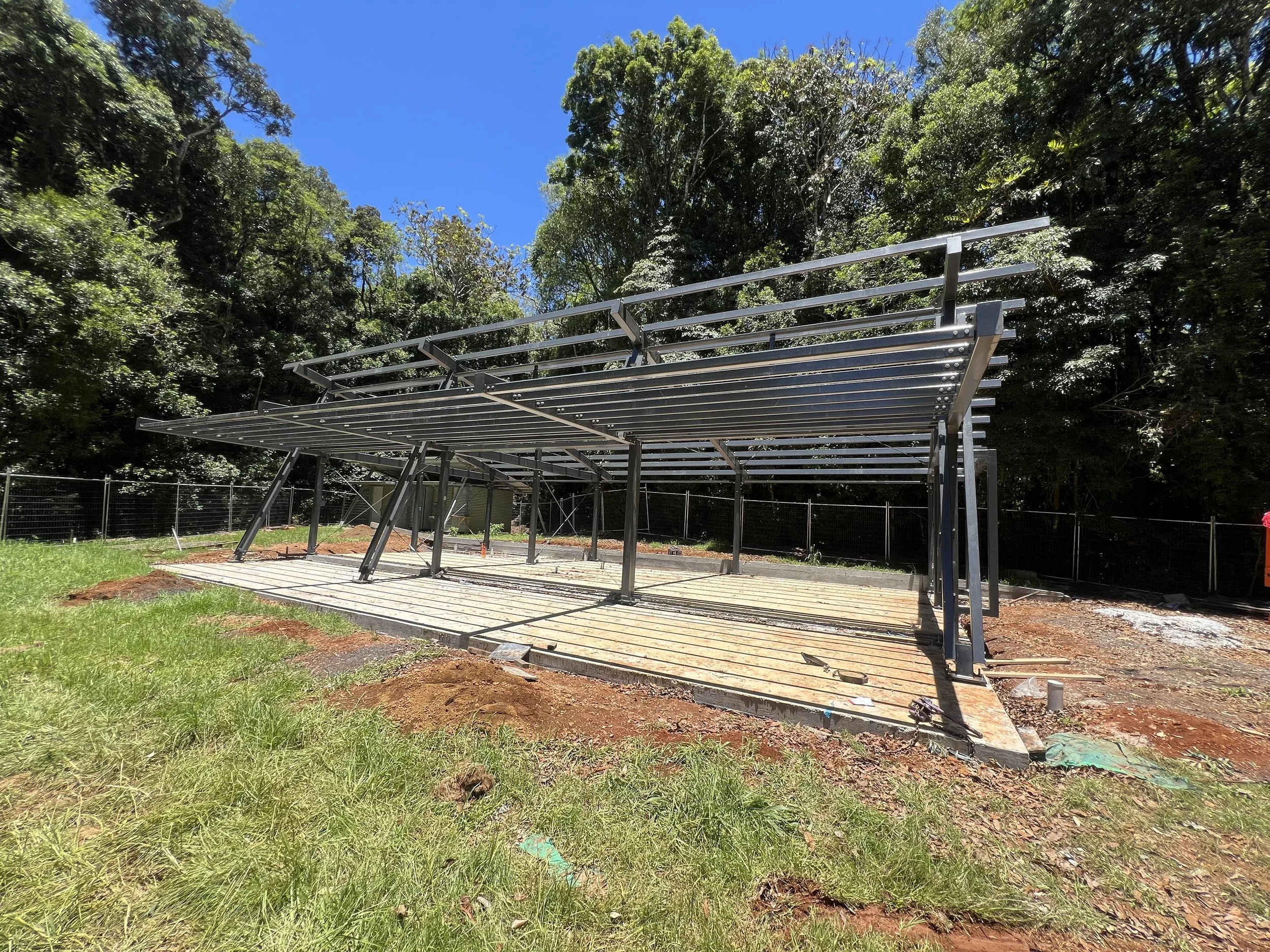Construction site with metal framework structure set on concrete foundation, surrounded by trees and a fence, under a blue sky.