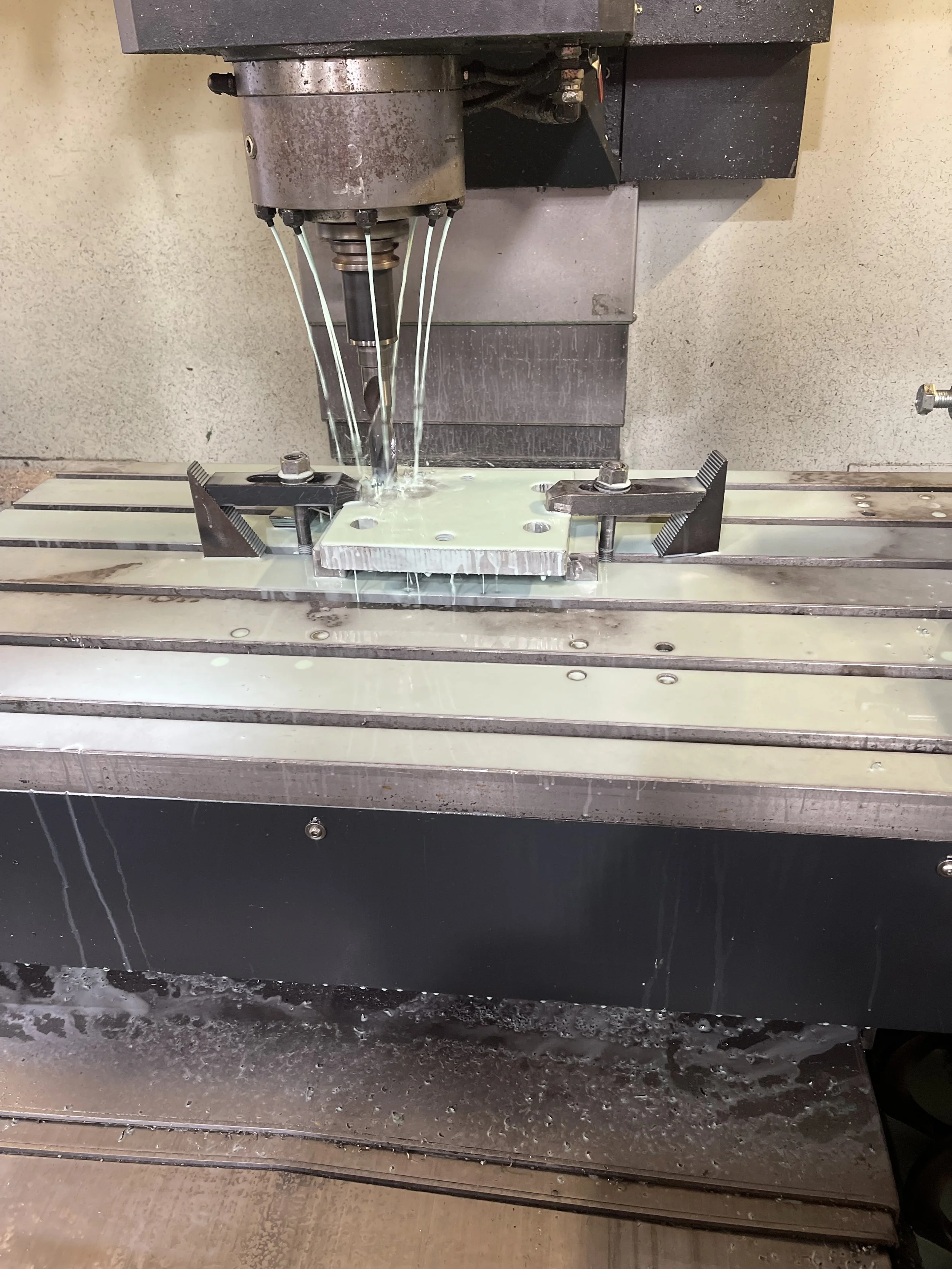 A metal CNC machine cutting a piece of metal on a worktable with metal clamps, with cutting coolant water flowing over the work area.