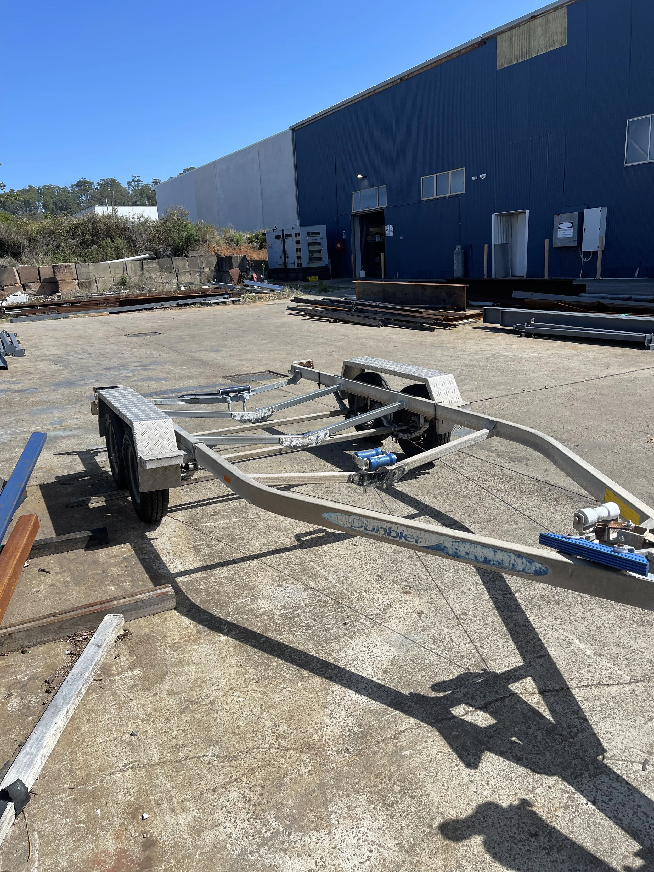 Empty boat trailer parked on concrete in front of a blue industrial building with some construction materials scattered around.