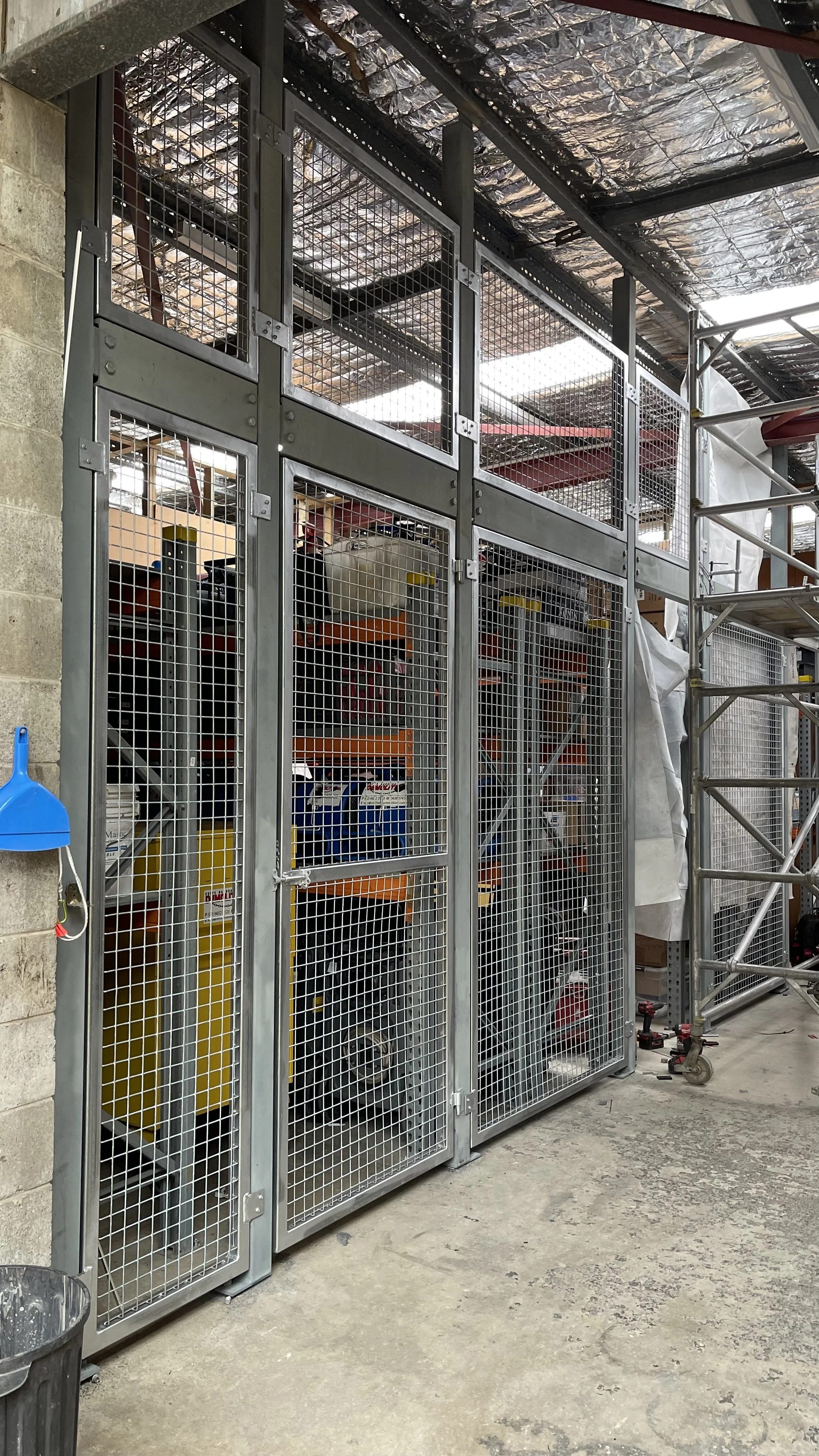 A metal storage cabinet with wire mesh doors containing various supplies, in a warehouse or workshop. There is a ladder on the right and a blue dustpan hanging on the left side.