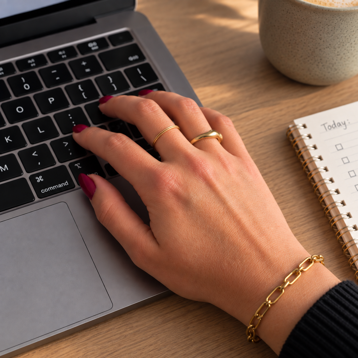 A hand with gold rings and a gold bracelet typing on a laptop keyboard on a wooden desk. A spiral notebook with checkboxes and 'Today:' written on it, a speckled coffee mug, and a black sleeve are also visible.