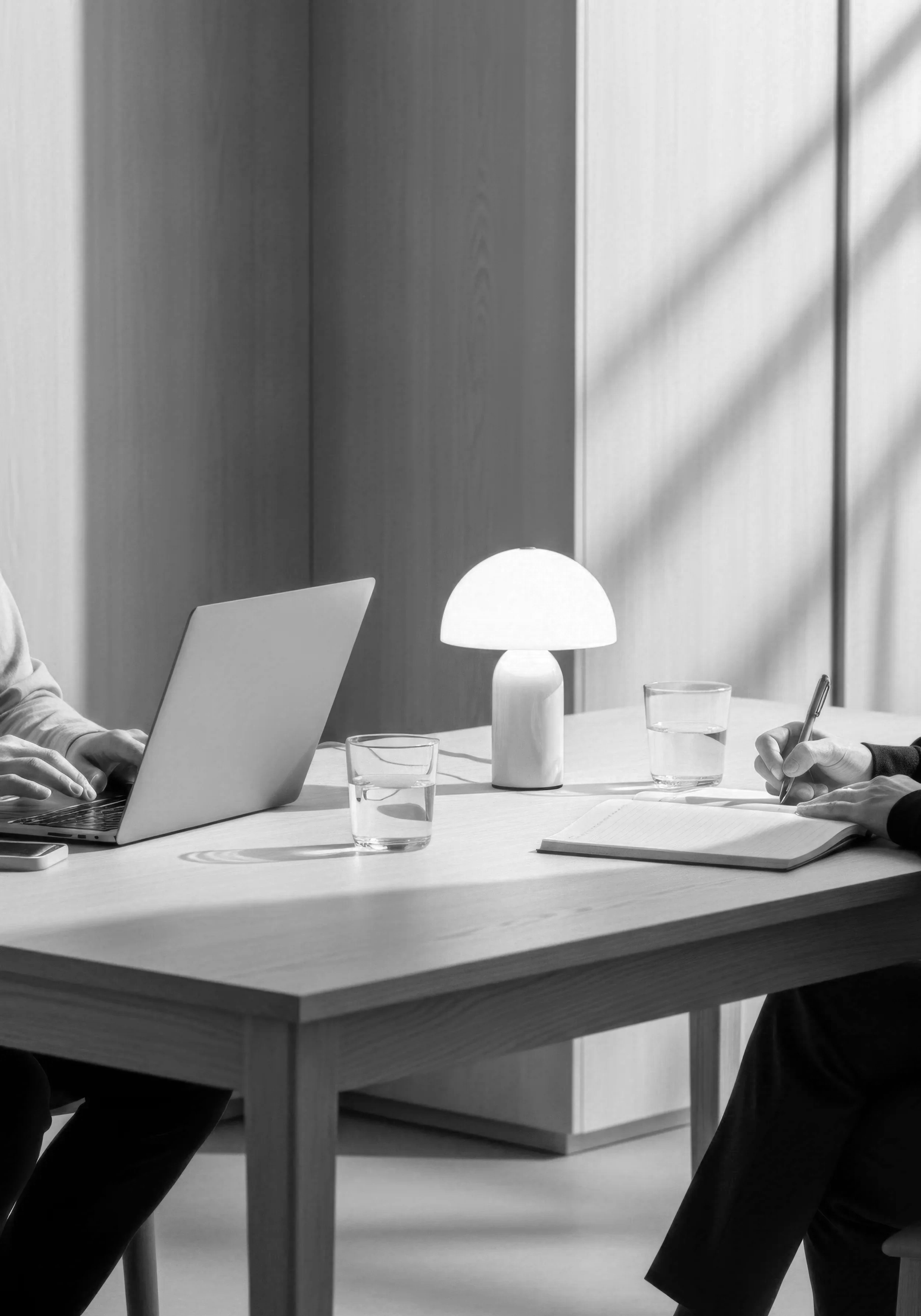 People sitting at a table working on laptops and writing in notebooks, with a lamp providing light, in a modern office setting.