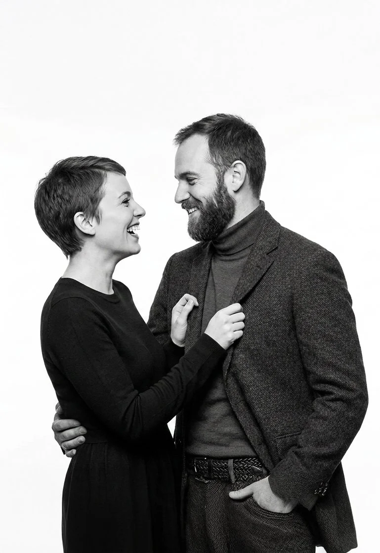 Black and white photo of a man and woman smiling and looking at each other closely. The woman has short hair and is wearing a long-sleeve shirt, while the man has a beard and is dressed in a blazer and turtleneck. They are holding each other affectio