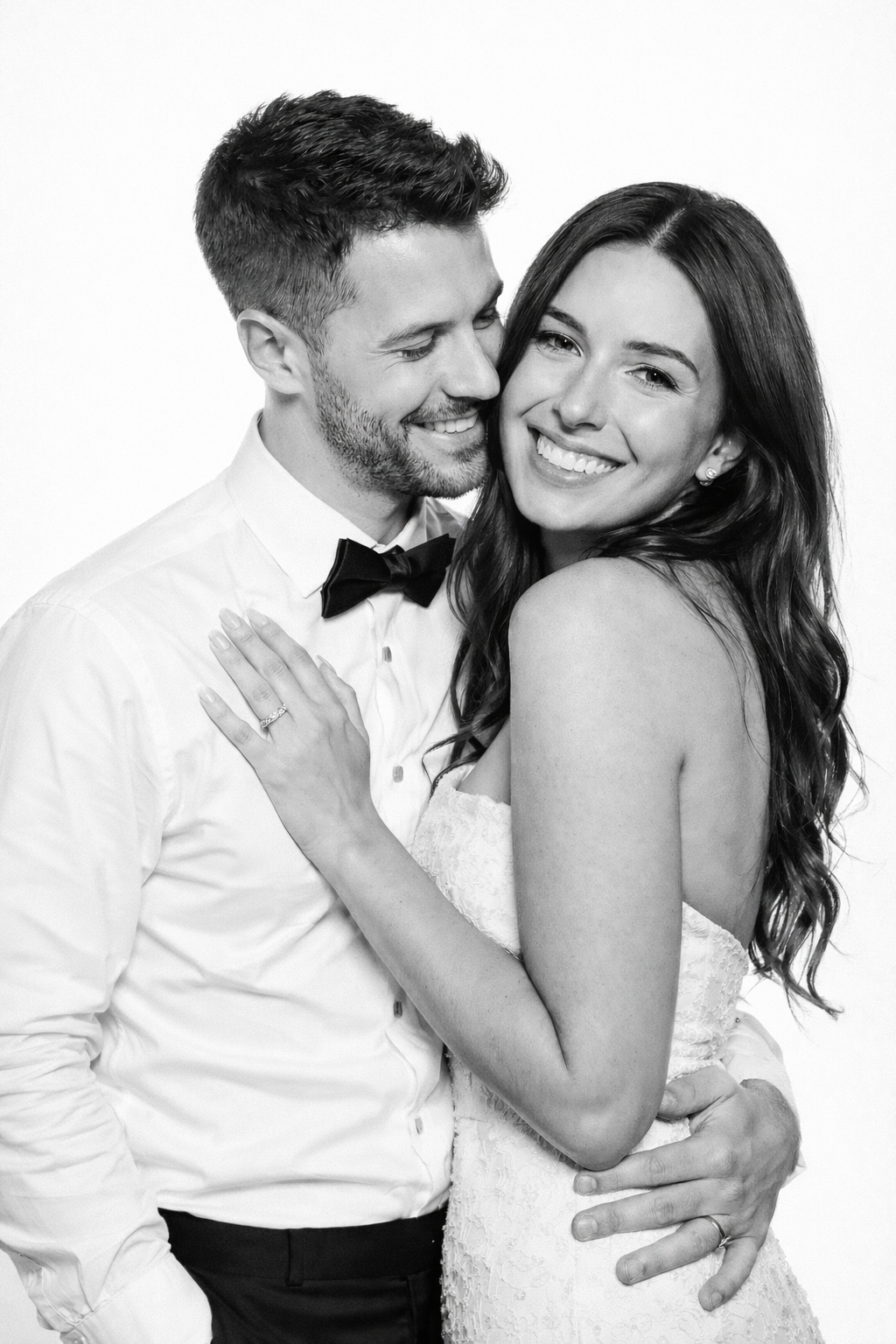 Black and white photo of a happy couple embracing, woman in lace wedding dress with long wavy hair smiling, man in tuxedo with bow tie smiling, woman showing engagement ring.
