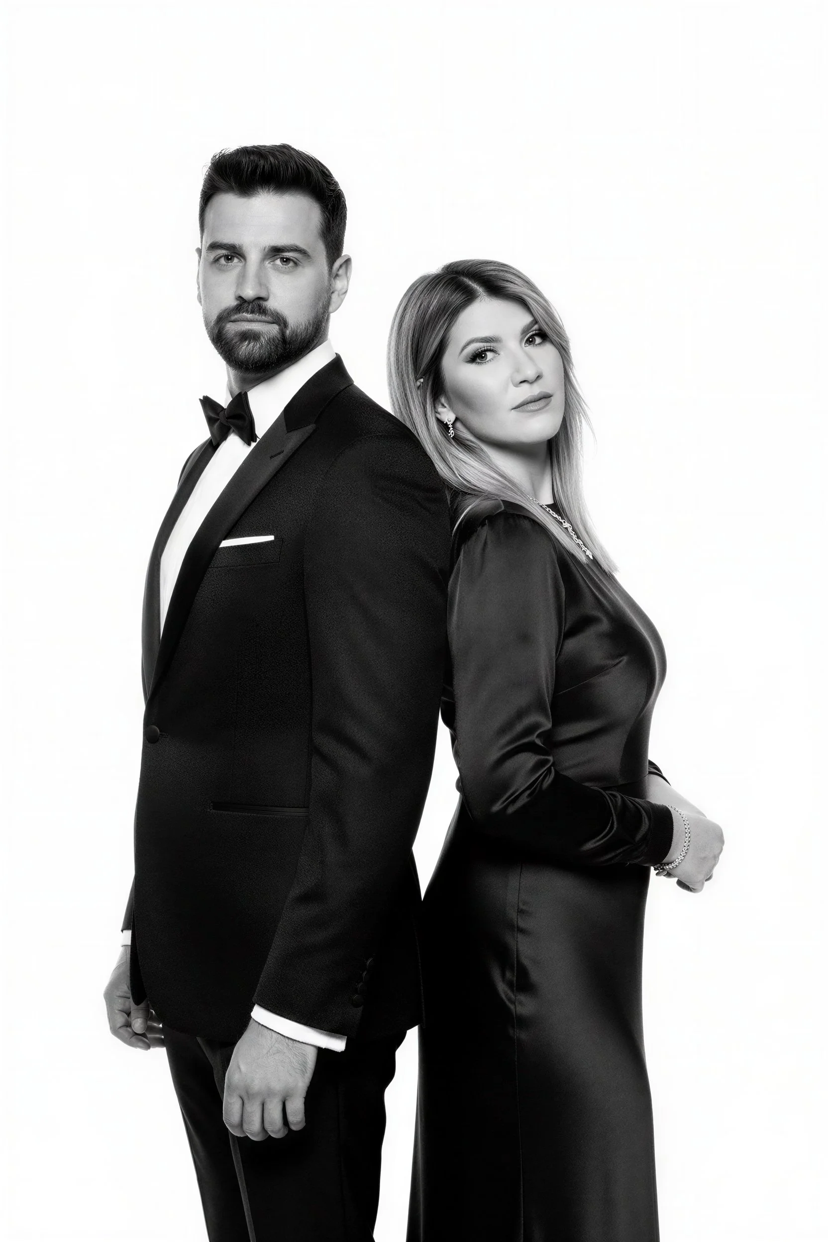 Black and white photo of a man and woman standing back to back, both dressed in tuxedo and elegant dress, looking serious at the camera.