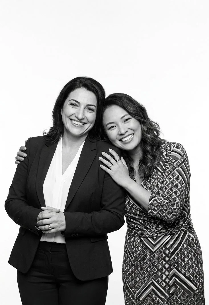 Two women smiling and embracing each other, one wearing a suit and the other in a patterned dress, in a studio setting with a plain background.
