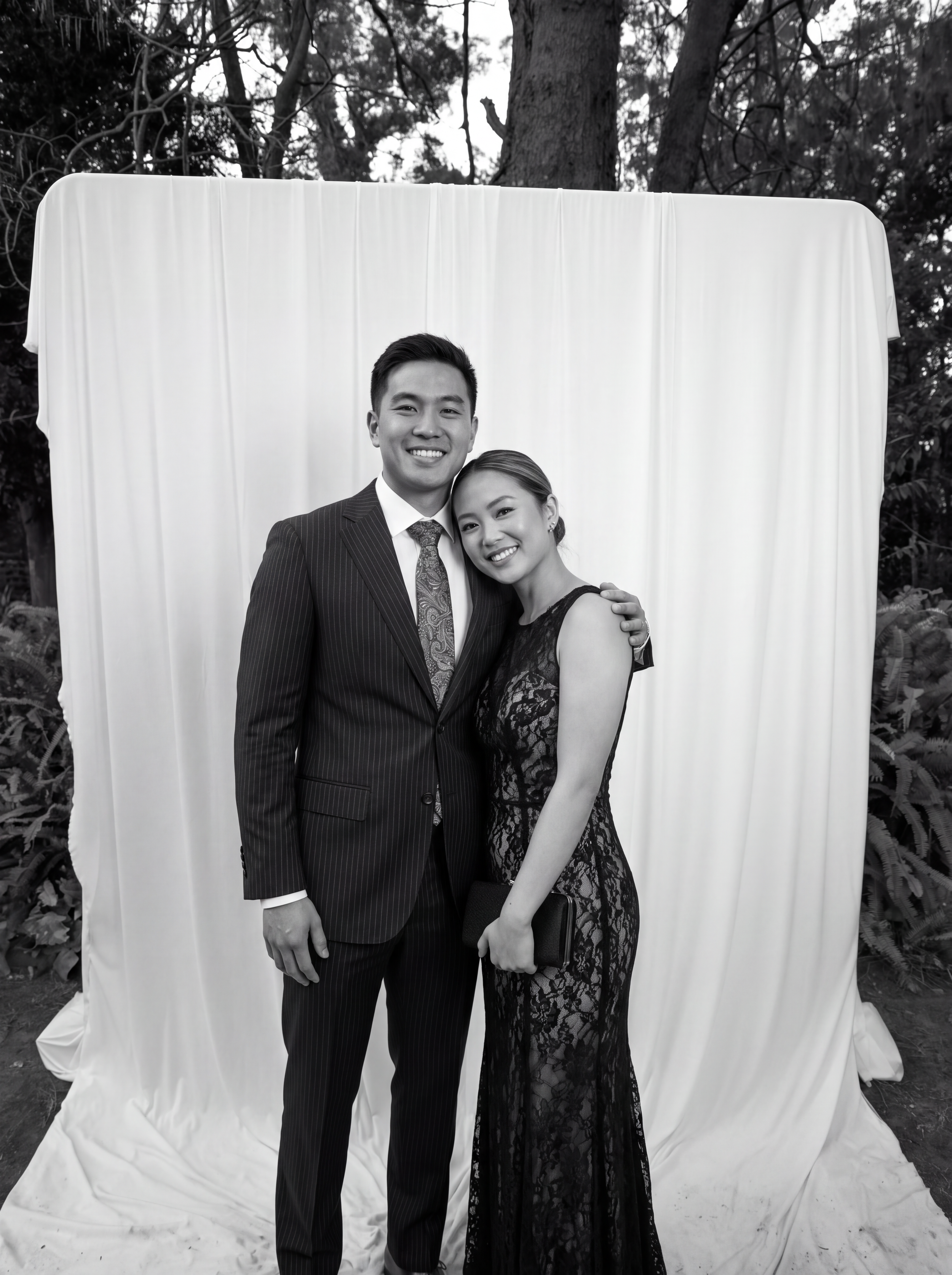 A smiling man in a suit and a smiling woman in a lace dress standing close together, holding a black clutch, in front of a white backdrop outdoors surrounded by trees.